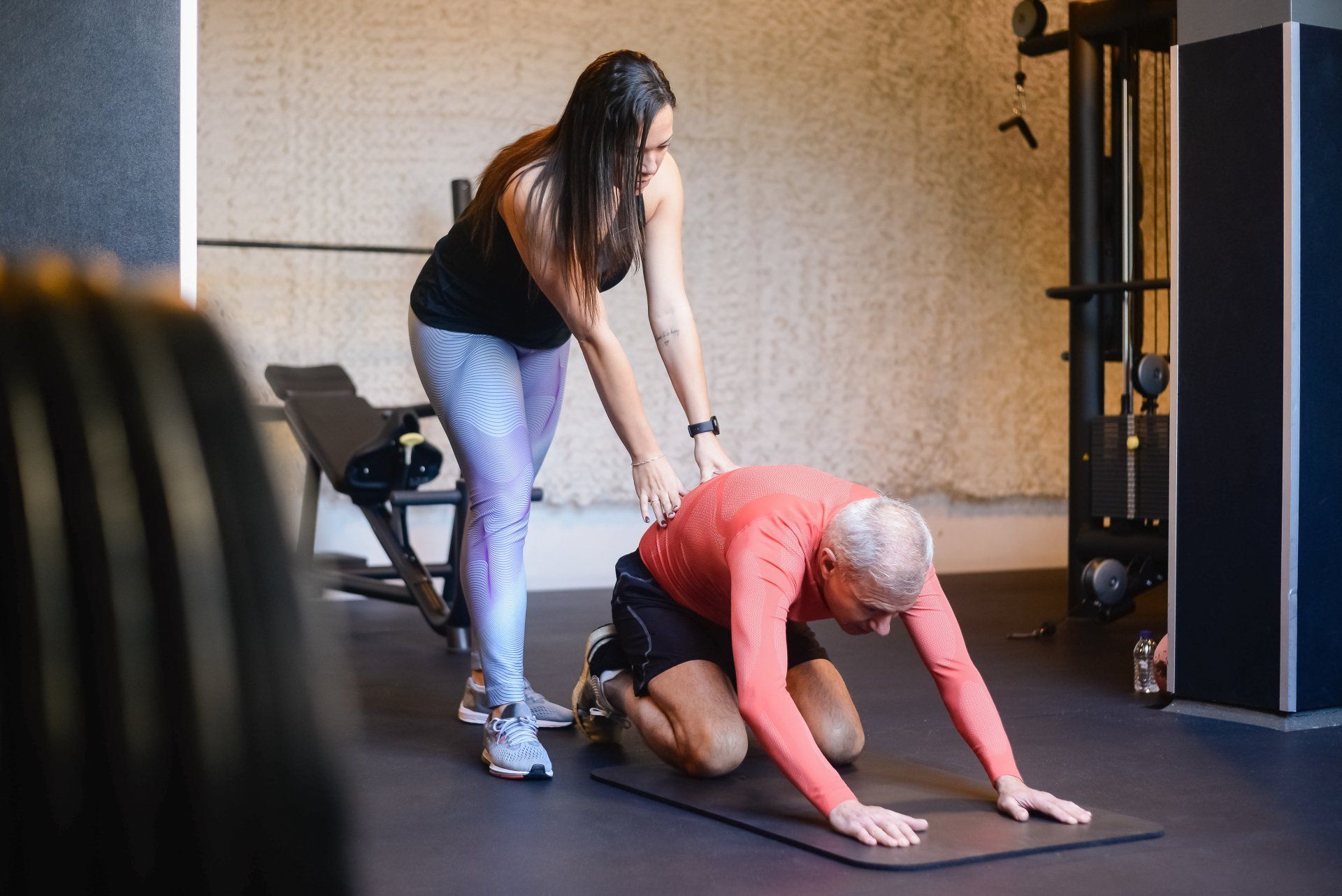 A woman is helping a man do stretching exercises on a yoga mat in a gym.