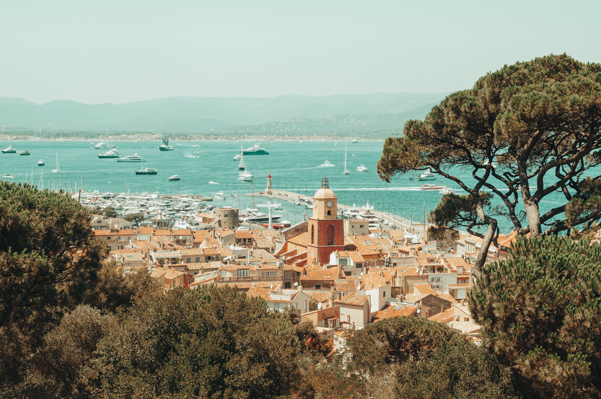 Vue d'une ville côtière aux toits de tuiles rouges, d'un port rempli de bateaux et d'une mer turquoise au loin.