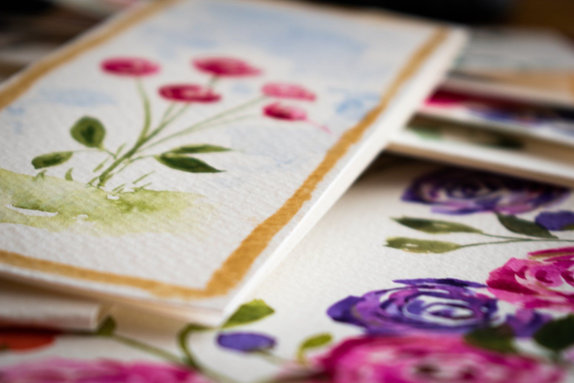 Watercolor floral artwork: close-up of pink, purple, and green flowers with gold borders on textured paper.