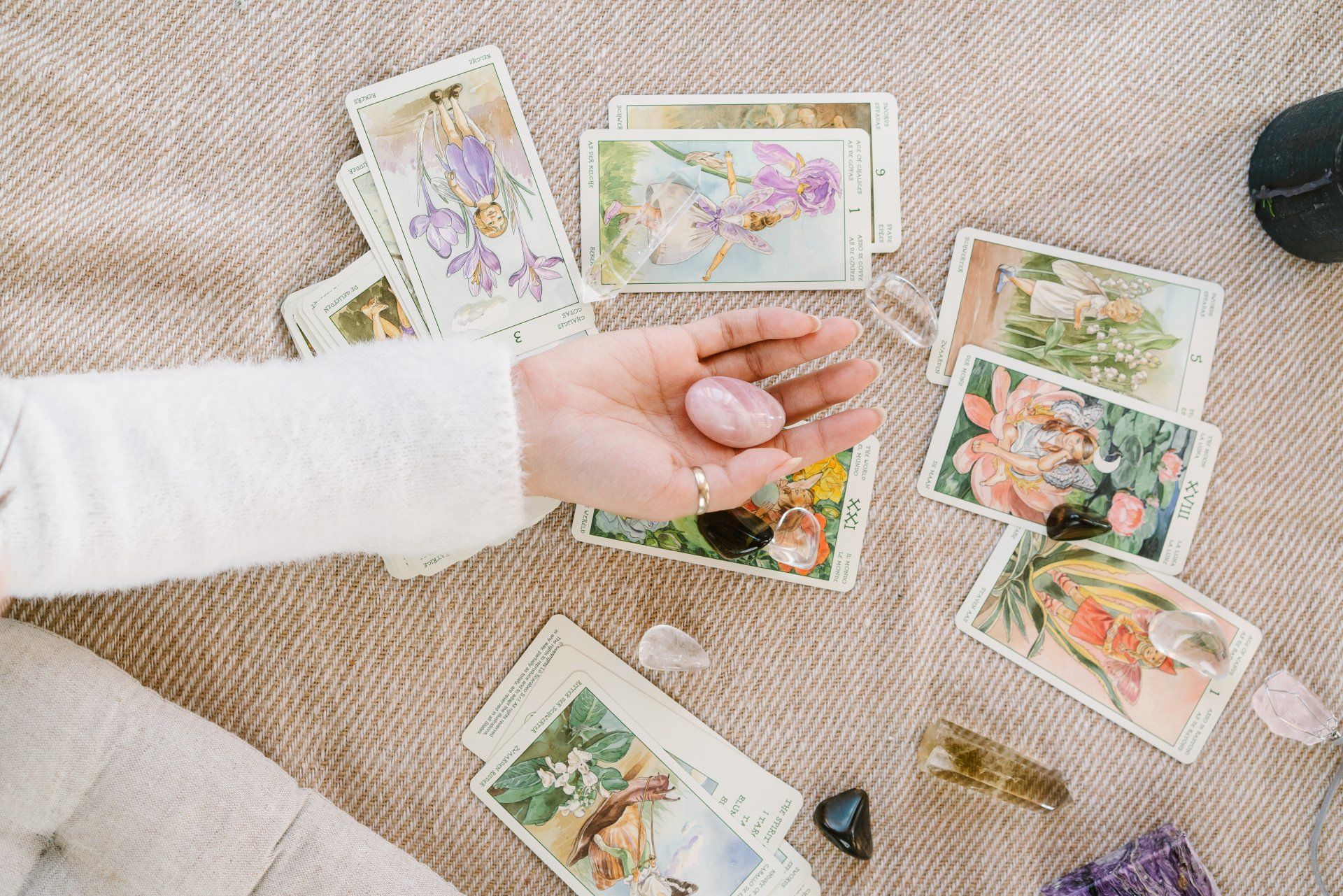 Een hand houdt een roze kristal vast, omringd door tarotkaarten op een beige ondergrond.