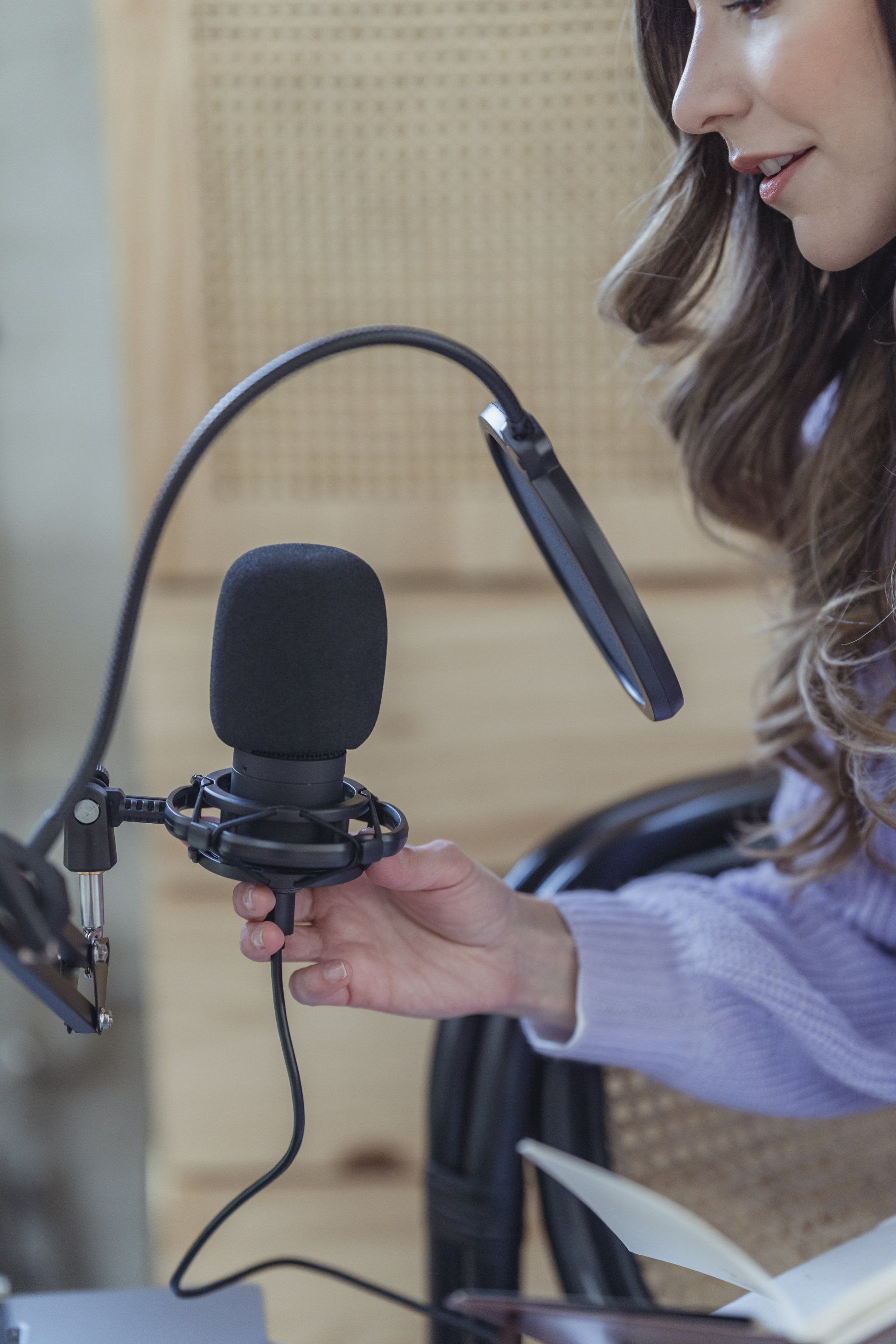 A person in a light purple sweater adjusts a studio microphone and pop filter while holding a notebook in an office setting.