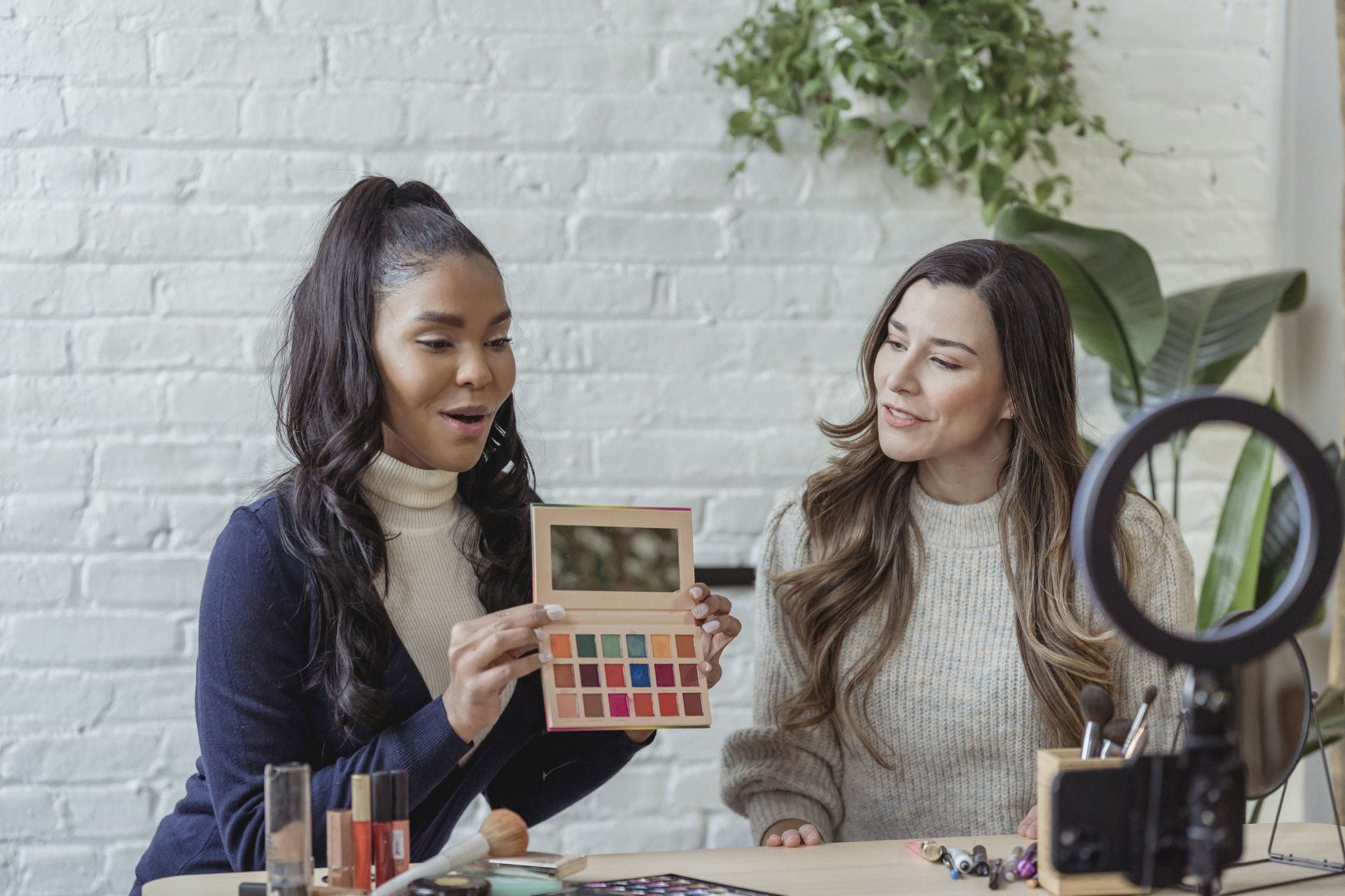 Two women at a table, demonstrating makeup. One holds a palette; both smile. Background with plants and a ring light.