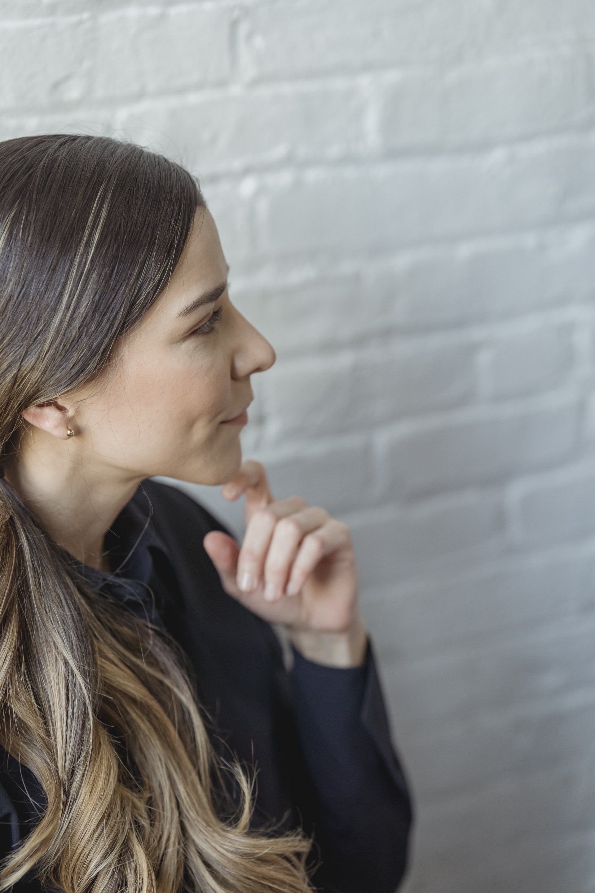A woman is sitting in front of a white brick wall with her hand on her chin.