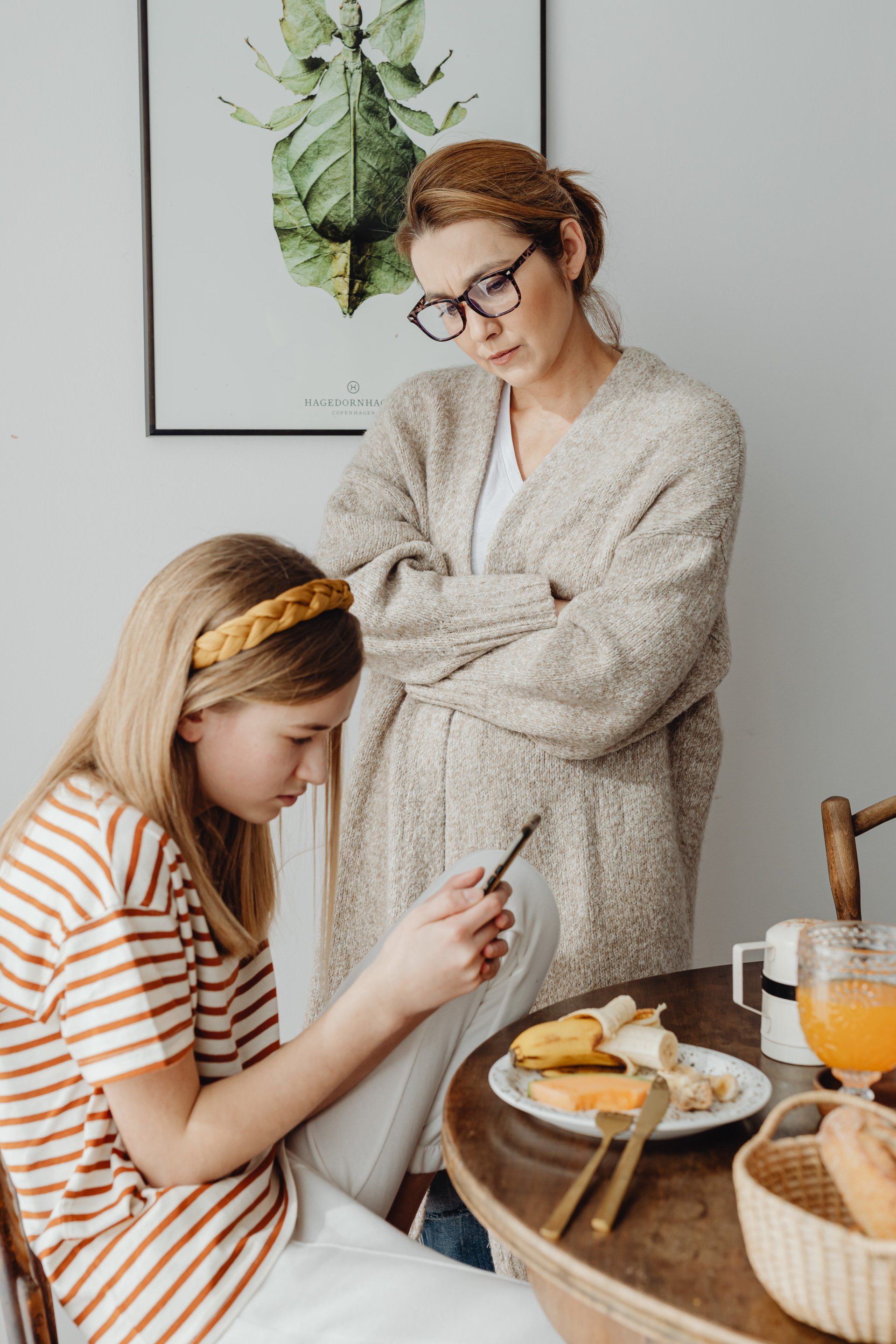 A woman is standing next to a girl sitting at a table looking at her phone.