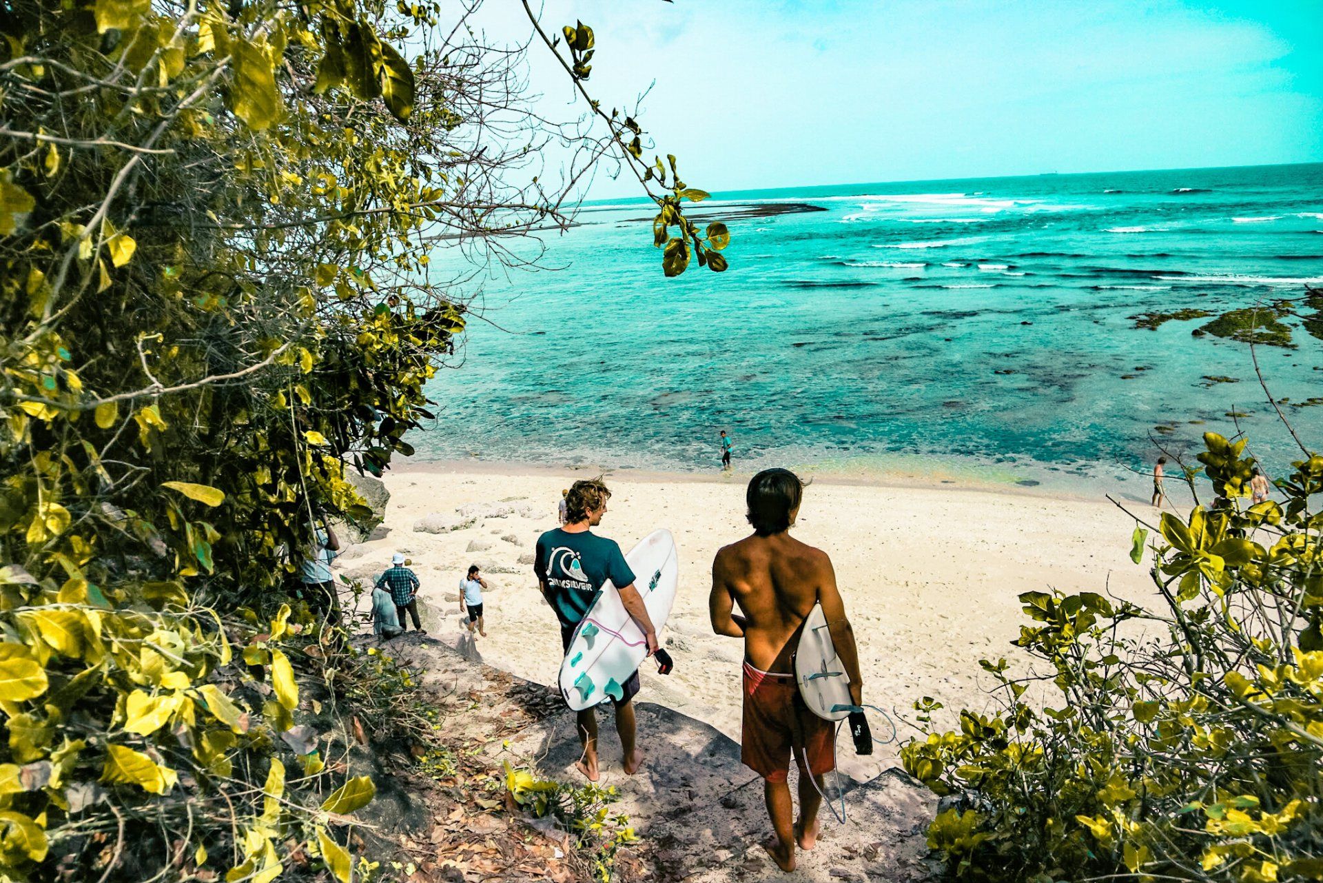 Two people are walking down a path to the beach carrying surfboards.