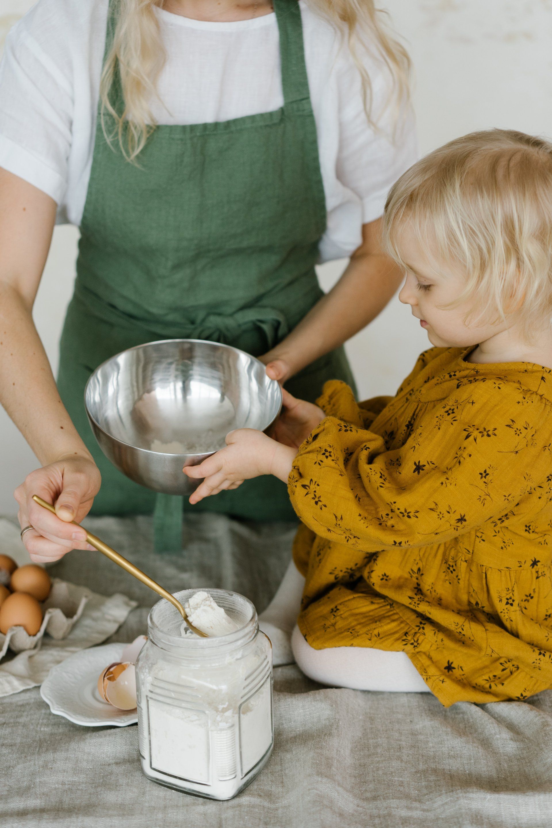 Woman in green apron and child in yellow shirt baking, scooping flour with spoon into a bowl.