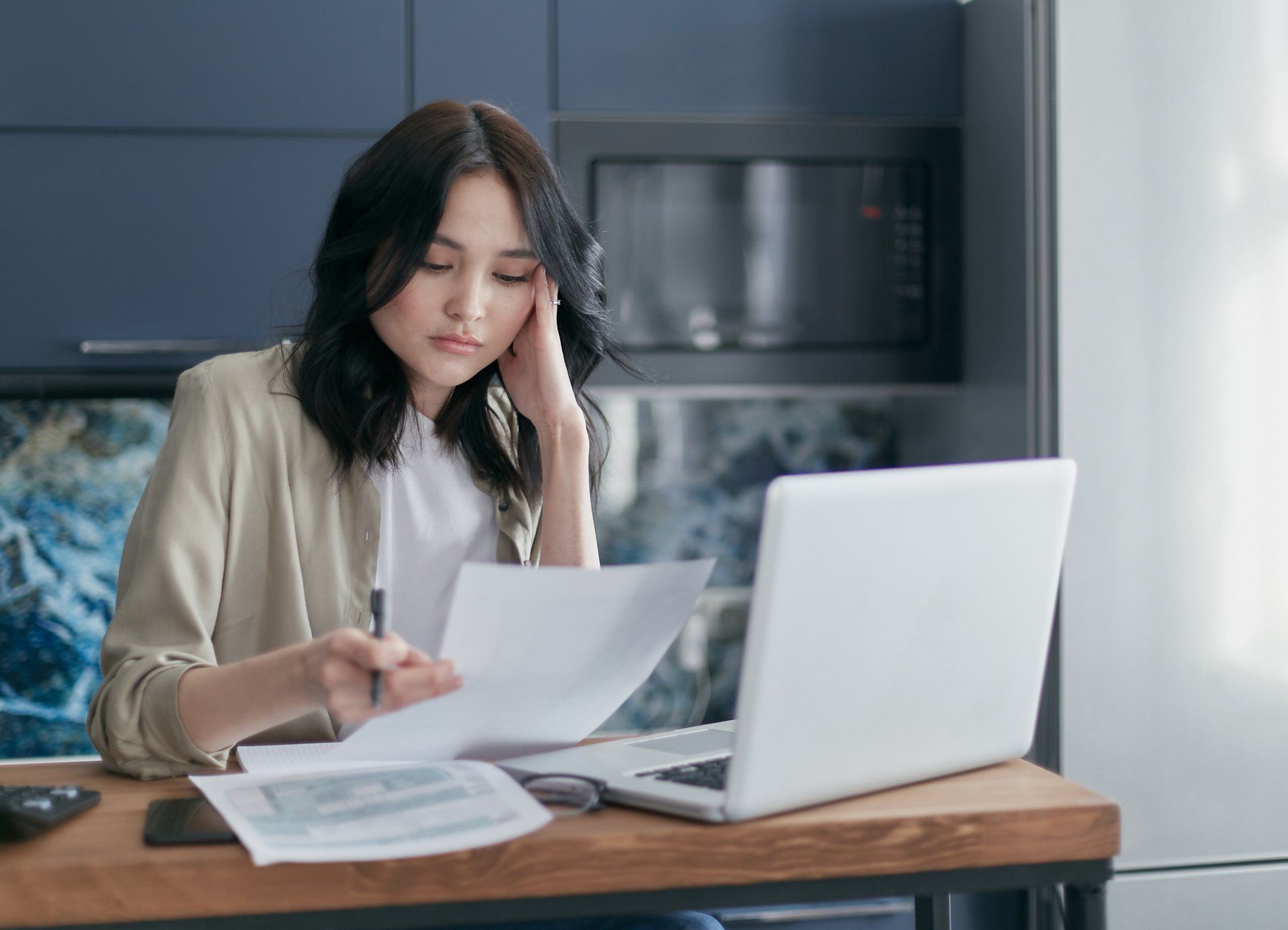 Woman, pensive, reviews documents and laptop at desk in kitchen.