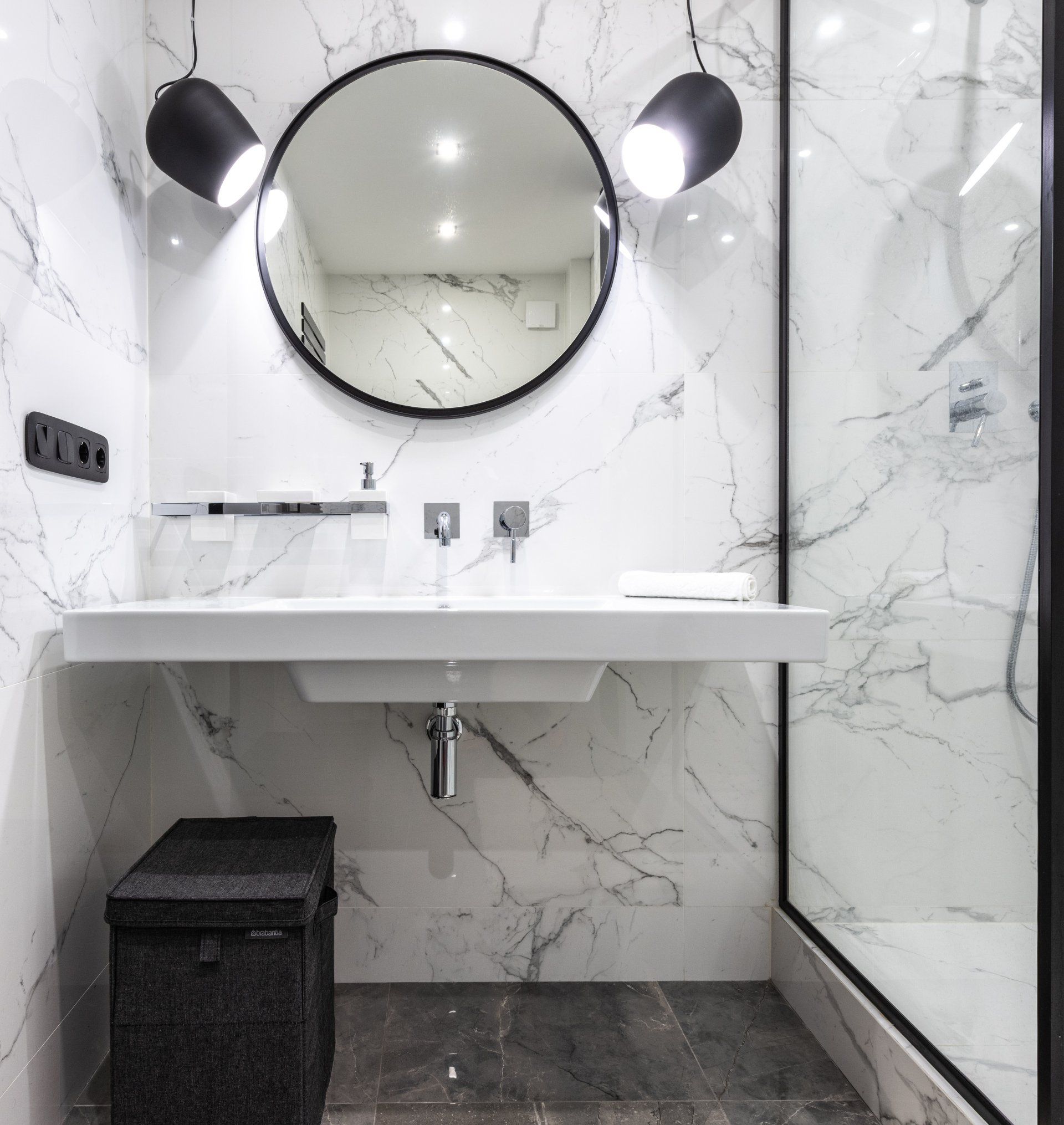 Salle de bains moderne avec murs en marbre, miroir rond, éclairage noir, lavabo blanc et rangements noirs.