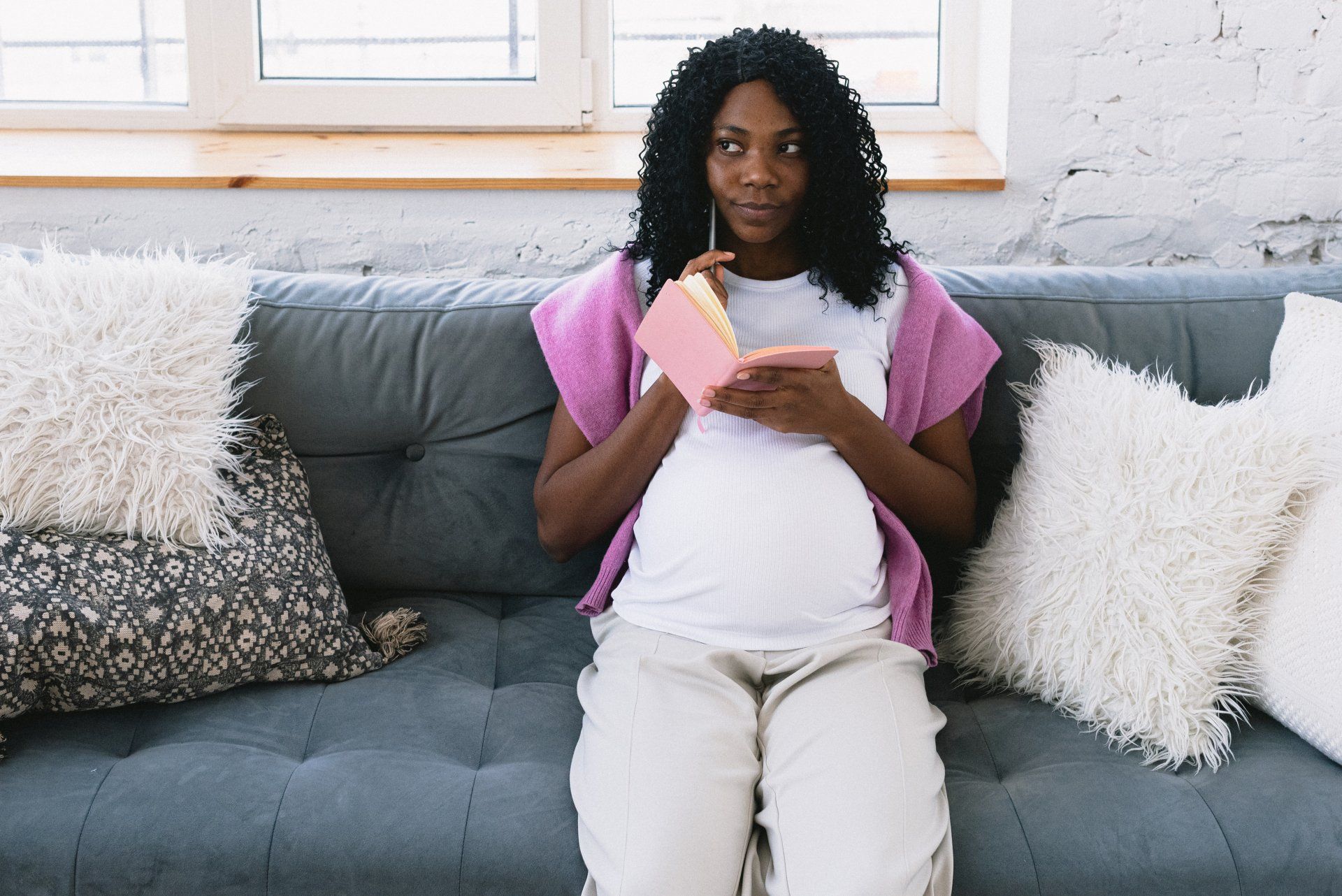 Pregnant person sitting on a couch, holding a pink cup, with pillows and a bright window behind them.