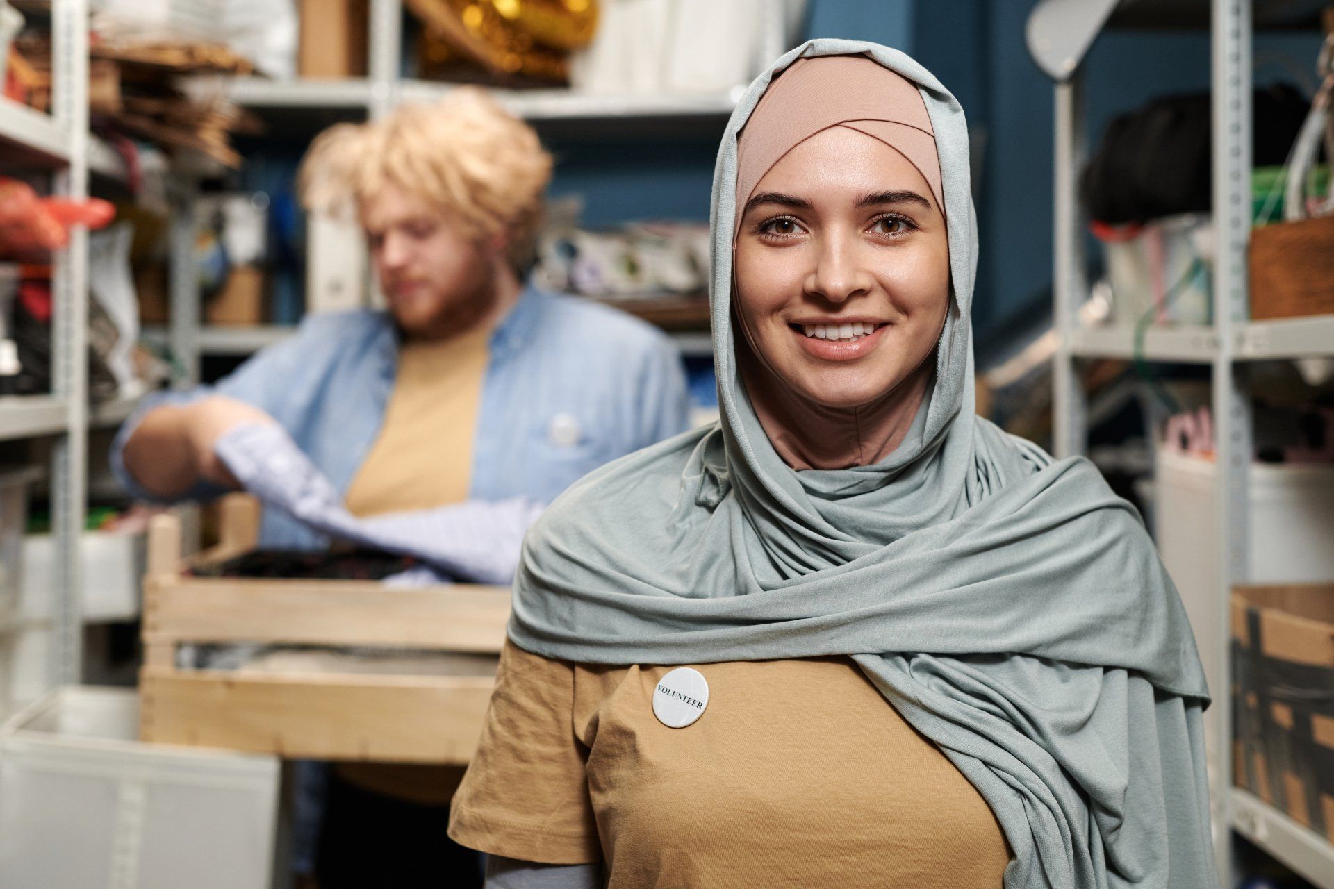 Woman in hijab smiling in a storage room, another person in background.