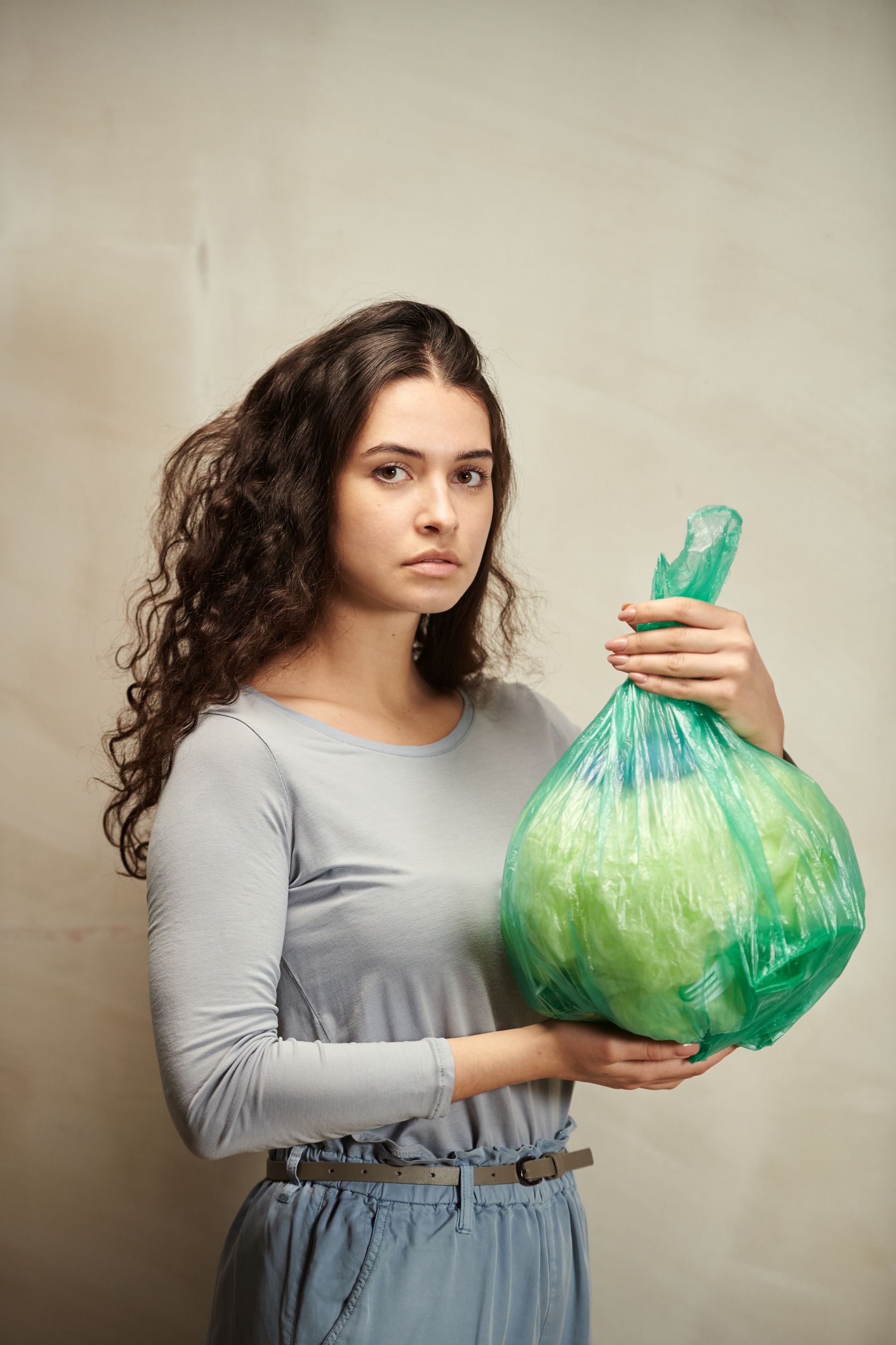 Woman holding a green plastic bag of green items against a neutral wall; she has curly hair and a serious expression.