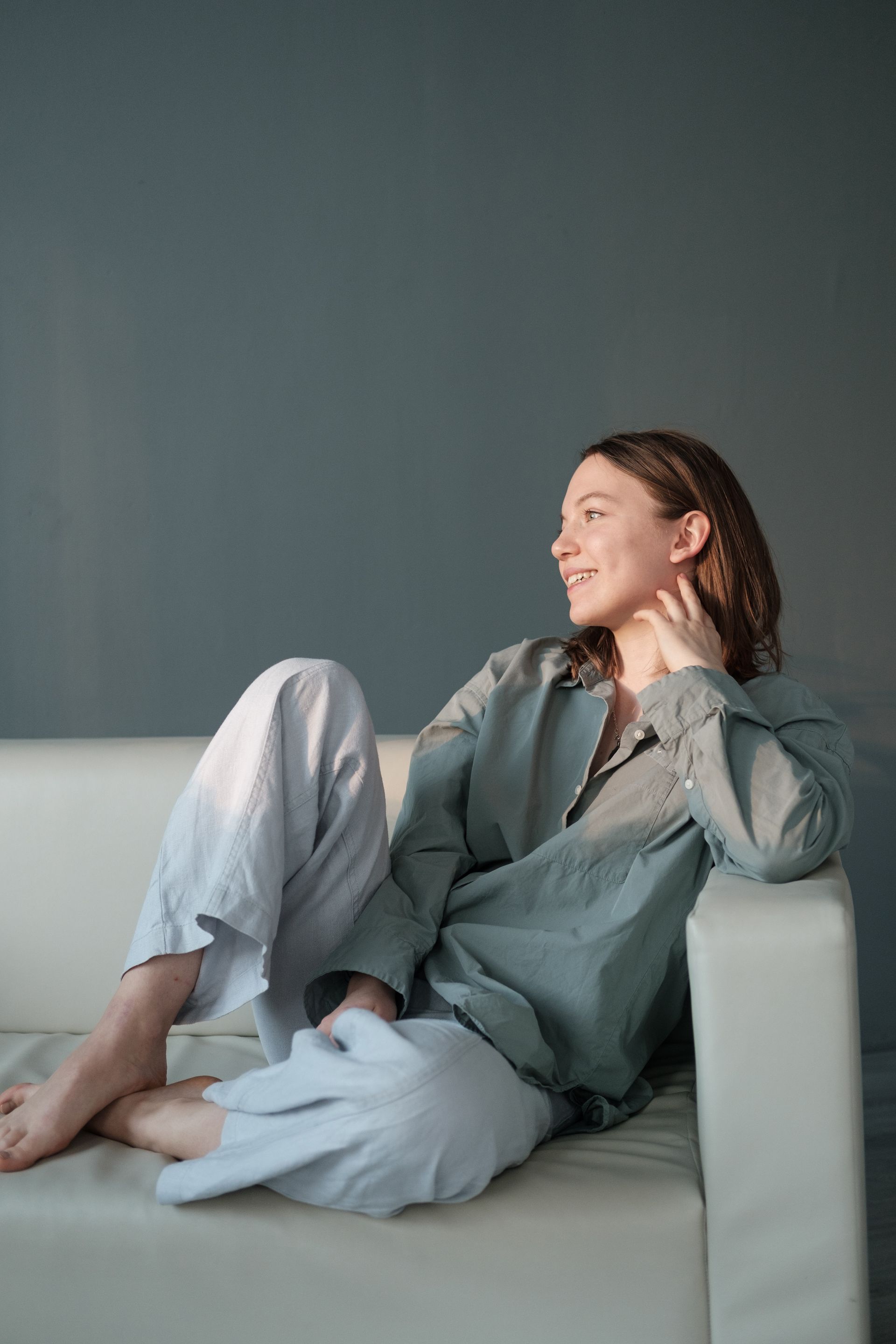 Woman reclining on a sofa, smiling in a gray outfit against a dark wall.