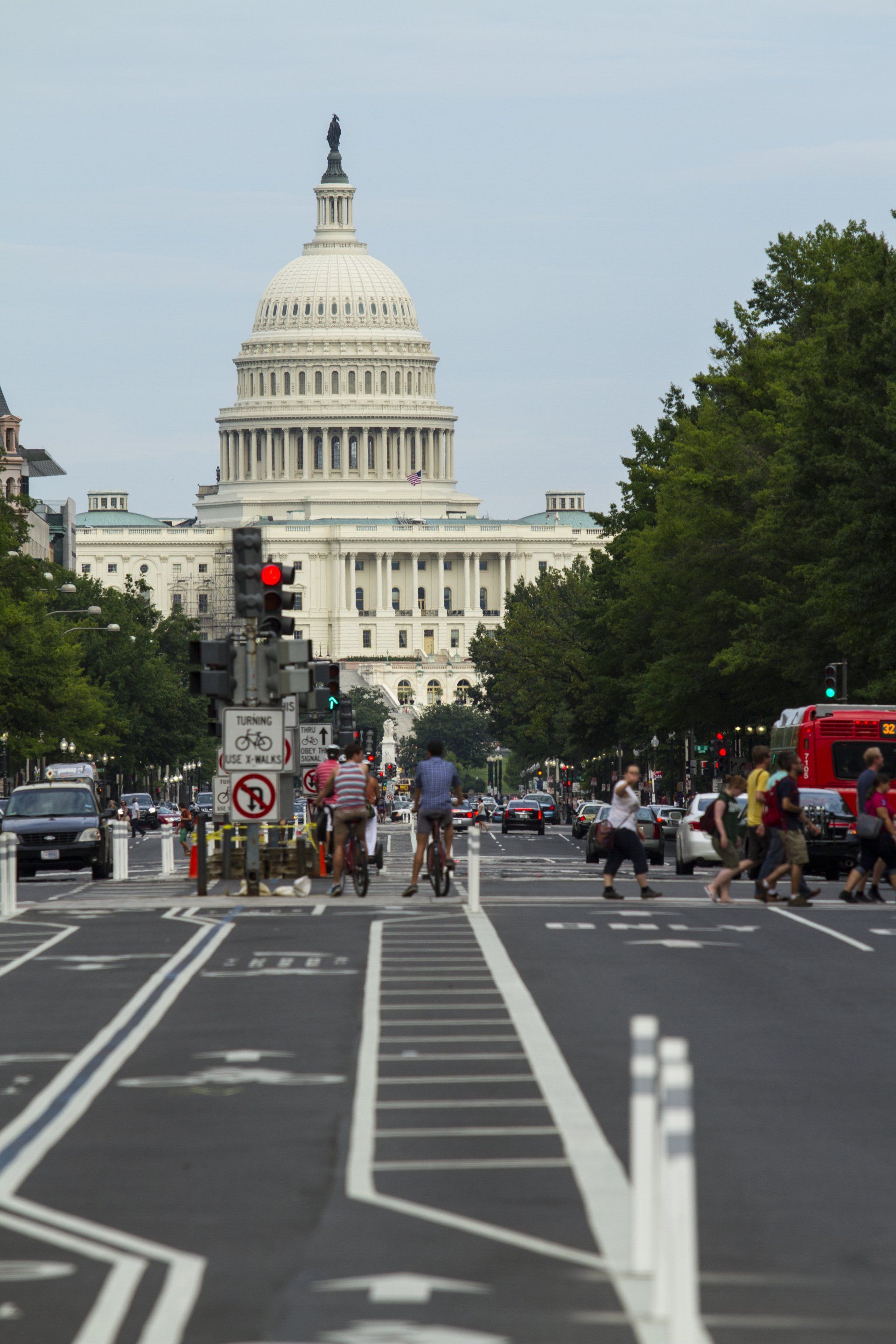 U.S. Capitol seen down a city street with pedestrians crossing and traffic, including a red bus.