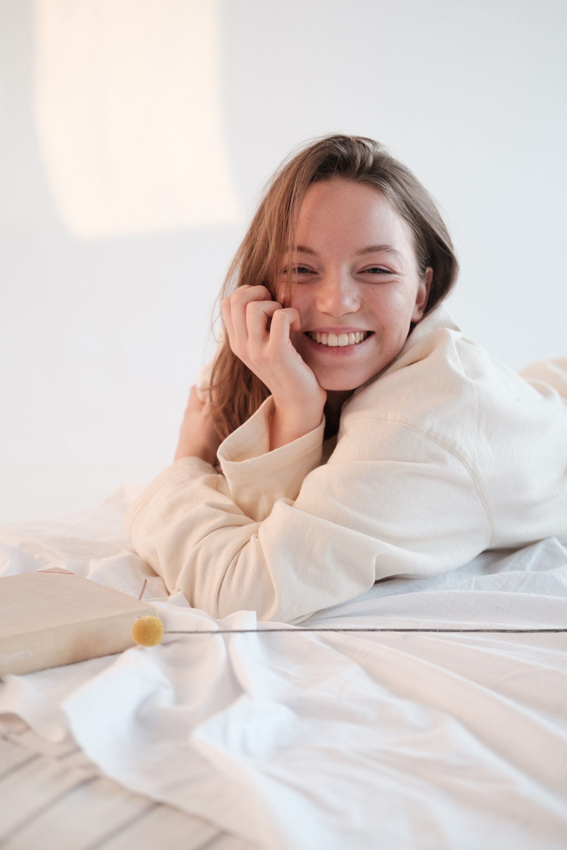 Woman smiling, resting on a white bed, wearing a light-colored top, hand to her face.