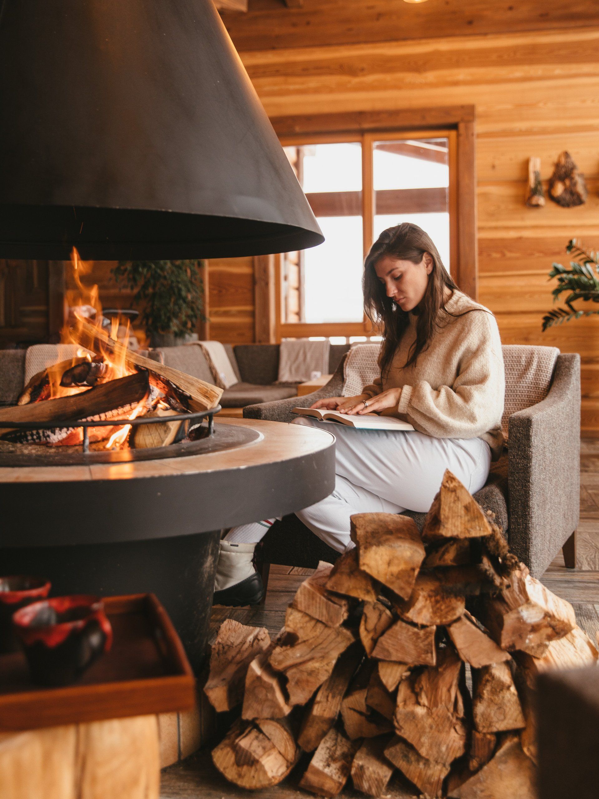 A person sits in a cozy wooden cabin reading a book next to a warm, glowing fireplace with a stack of firewood nearby.