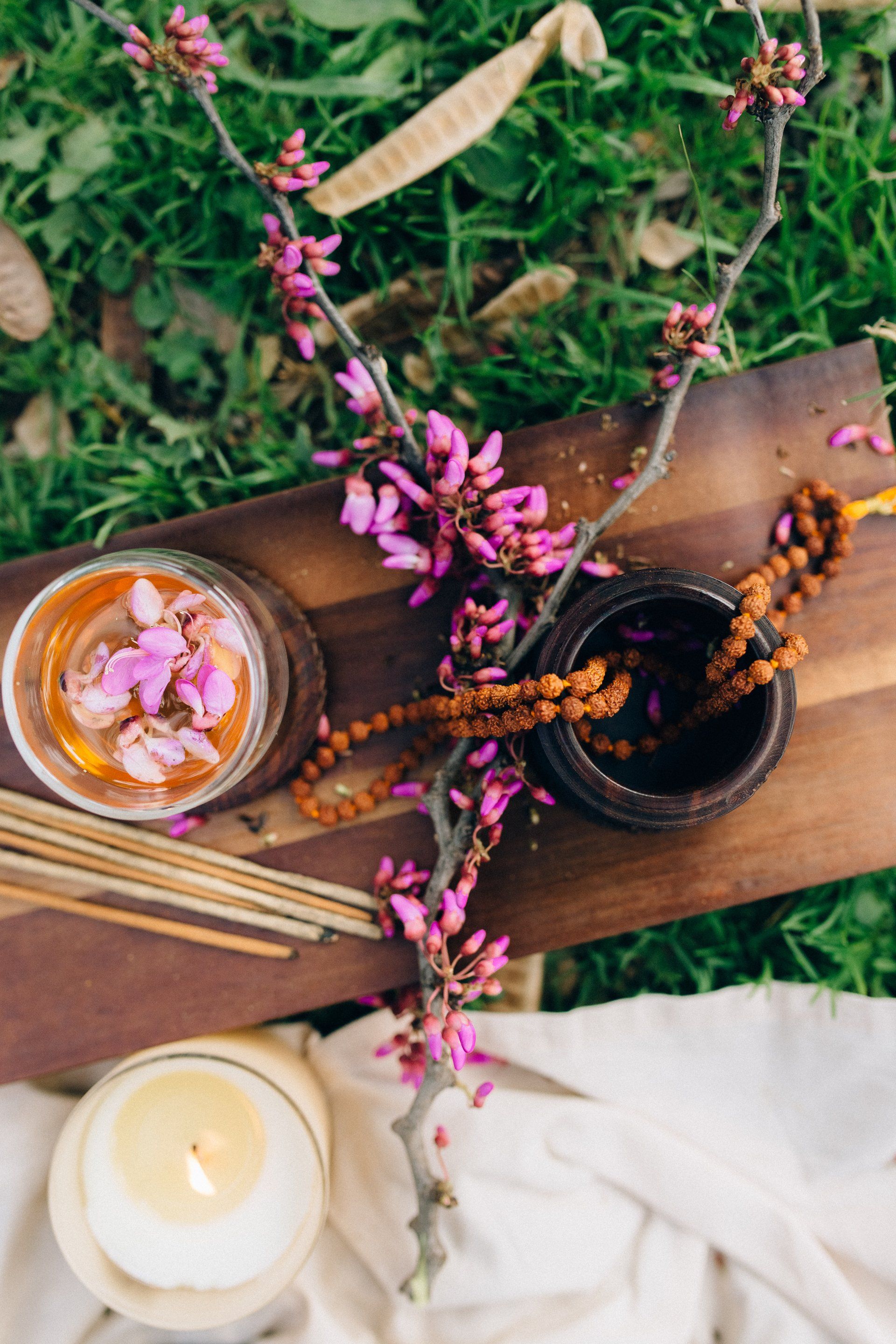 Wooden tray with drink, flowers, beads, candle, and incense on grass.