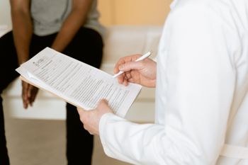 A doctor is writing on a clipboard while a patient sits in the background.