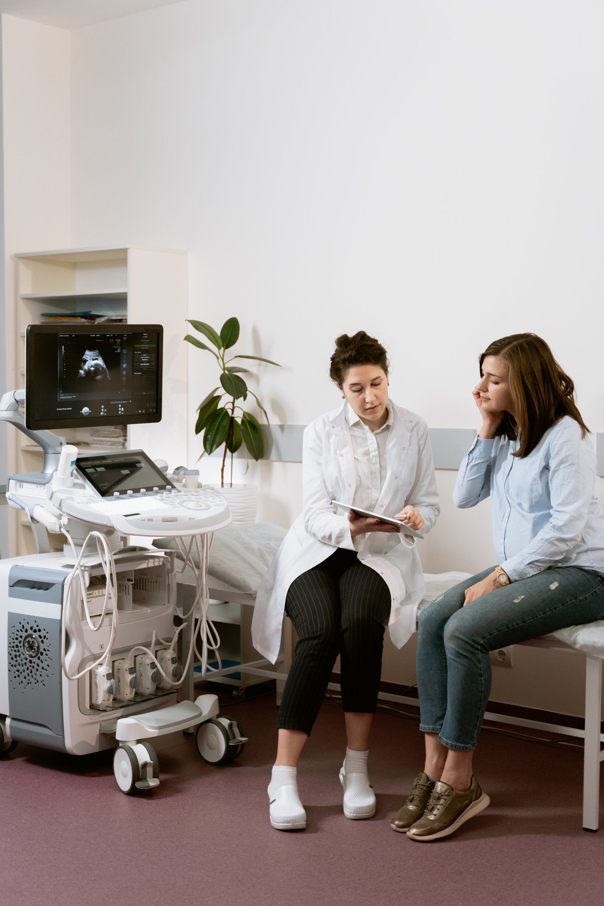 Two women are sitting on a bench in a waiting room in front of an ultrasound machine.