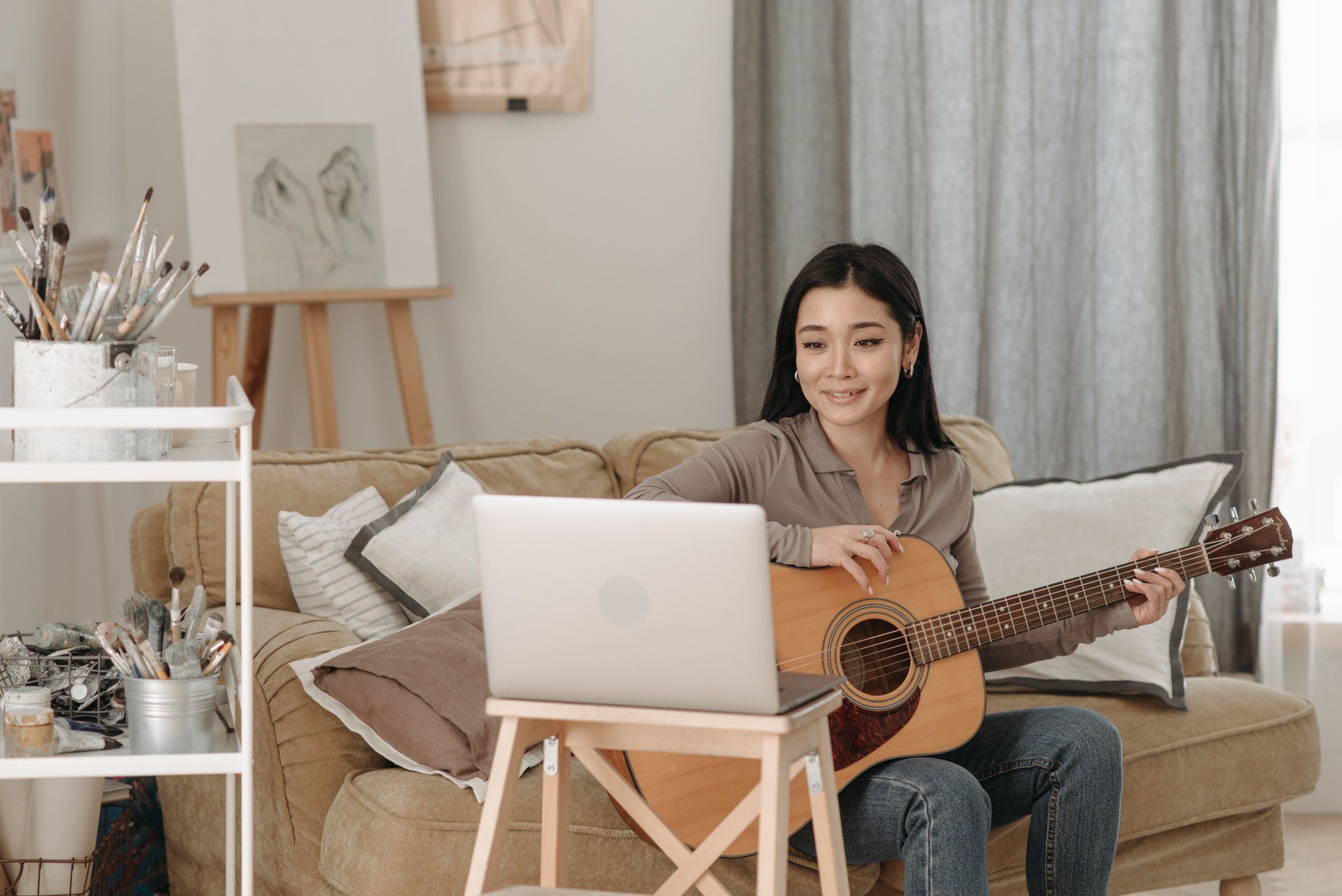 Woman plays acoustic guitar in front of a laptop, smiling, on a couch. Living room setting.