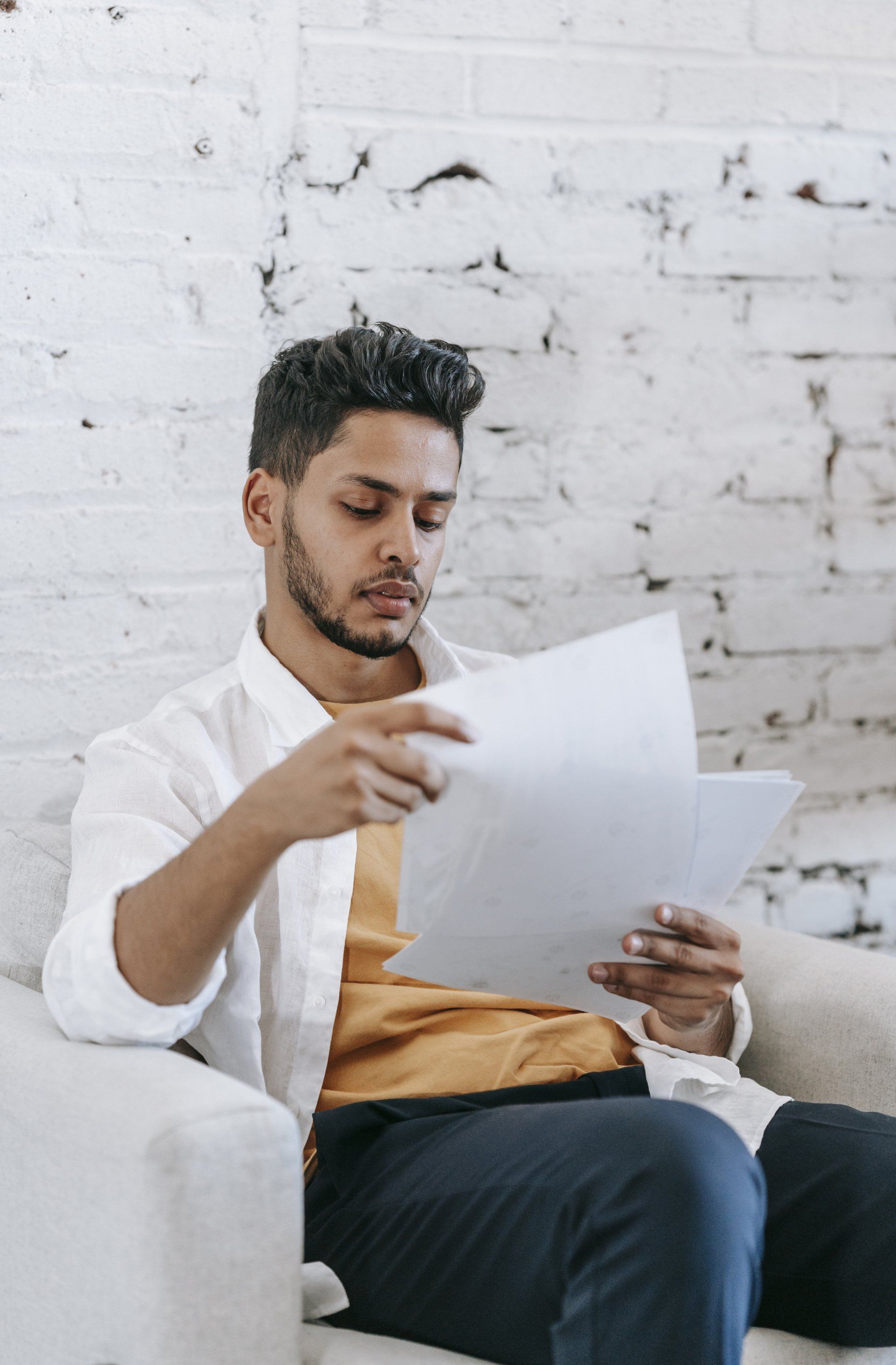  Man in white shirt looking at papers, seated on a white chair, white brick wall background.