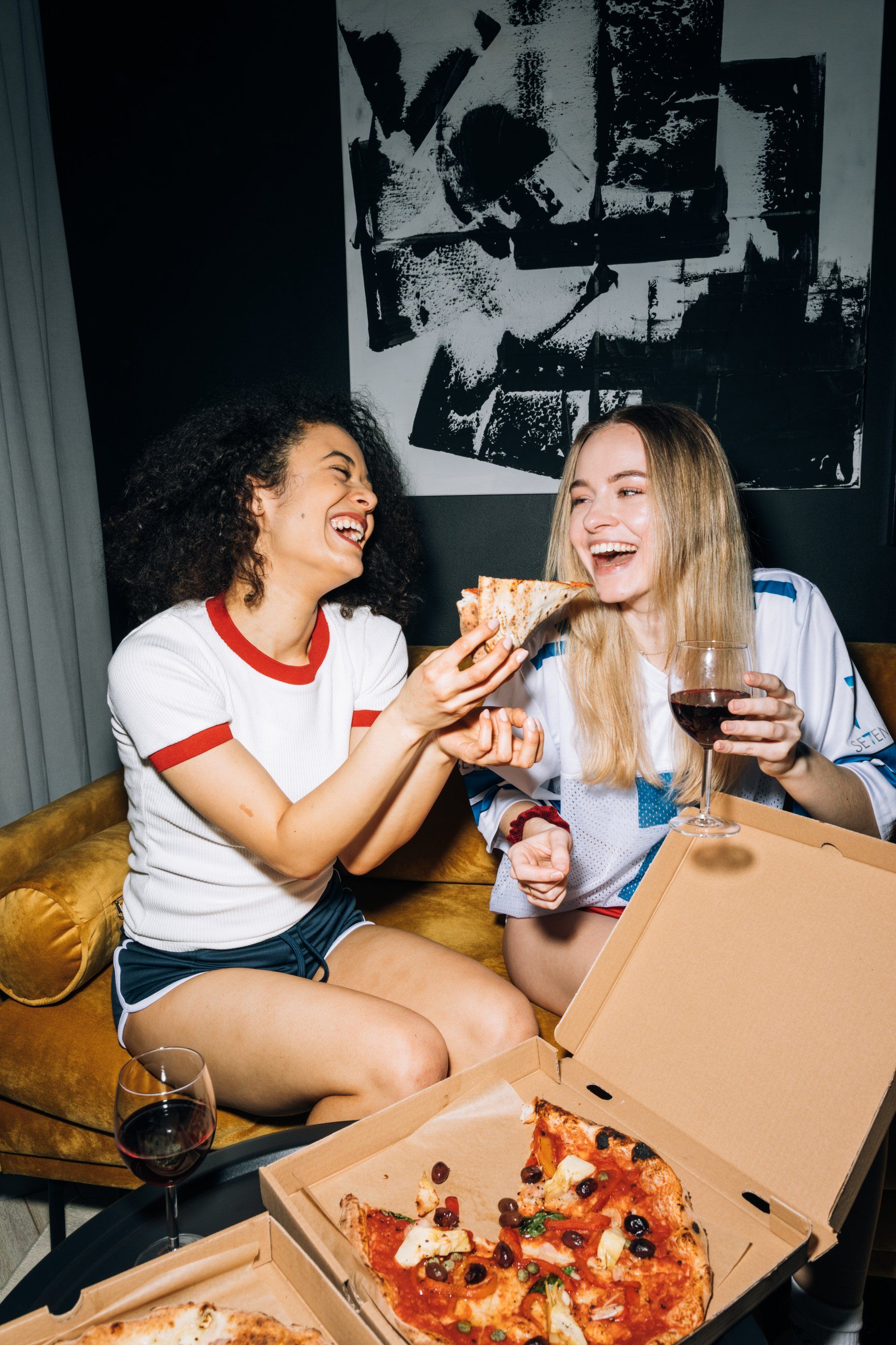 Two women are sitting on a couch eating pizza and drinking wine.
