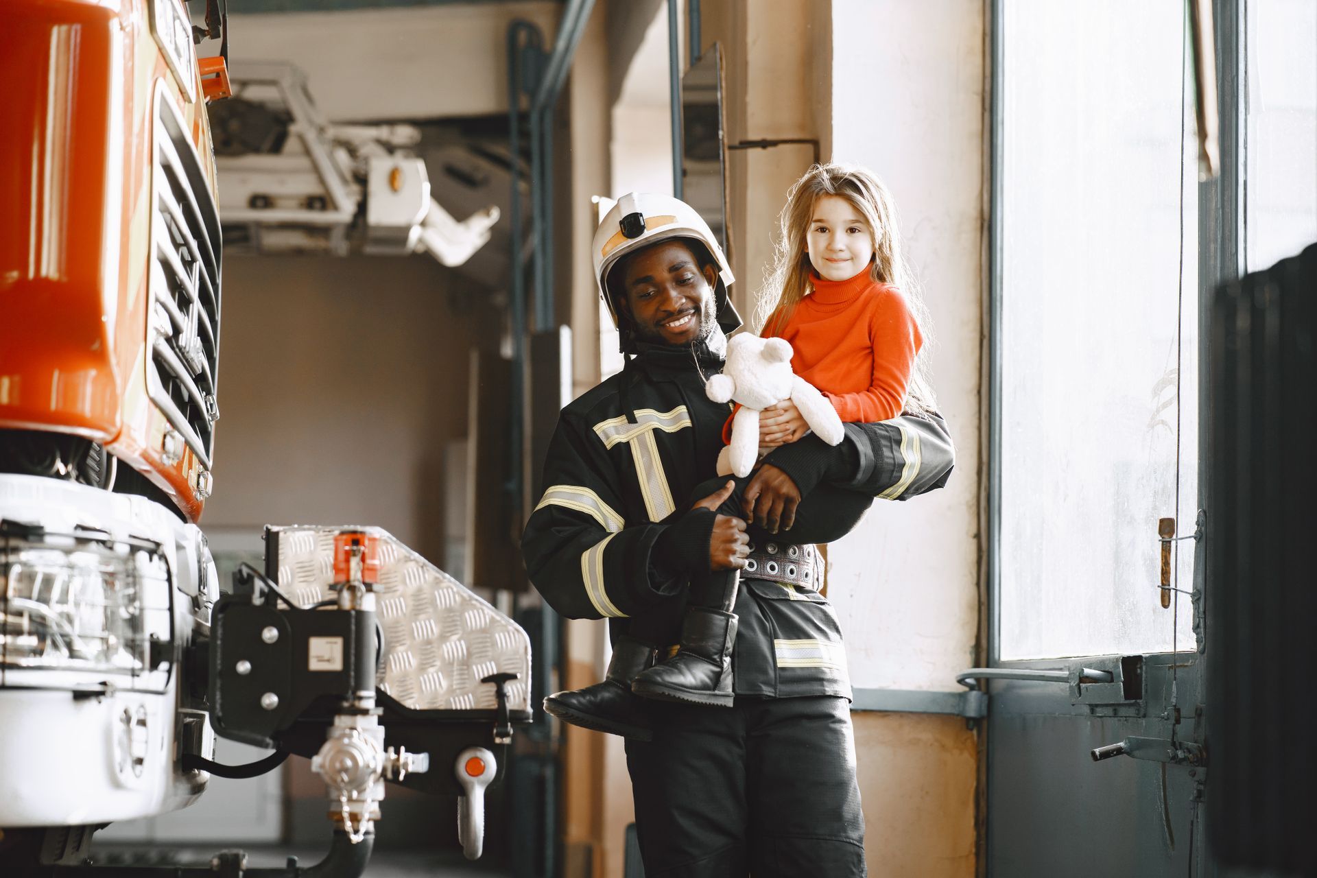 Firefighter holding a child, both smiling, near a fire truck inside a station.