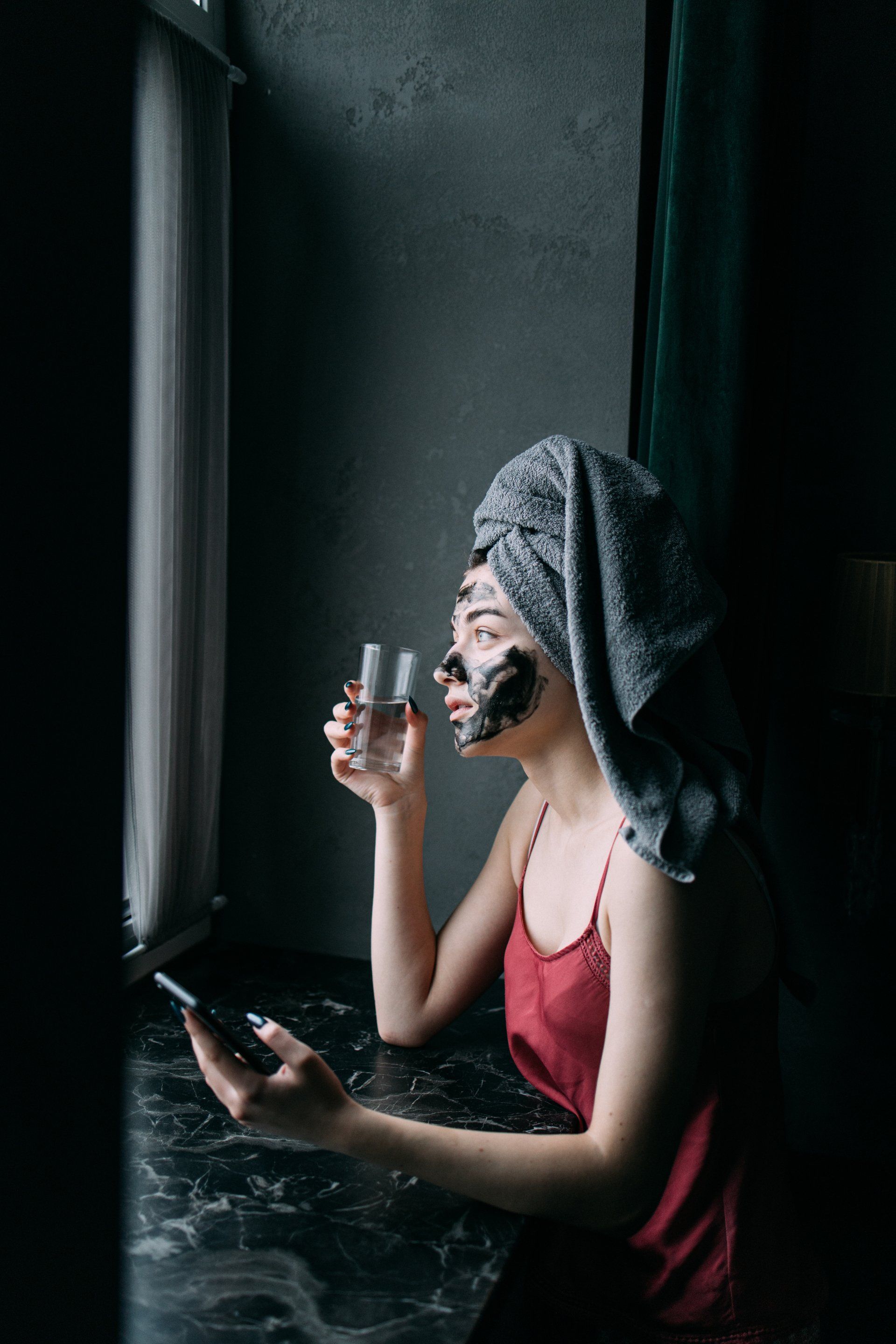 A woman with a towel wrapped around her head is drinking water and looking at her phone.