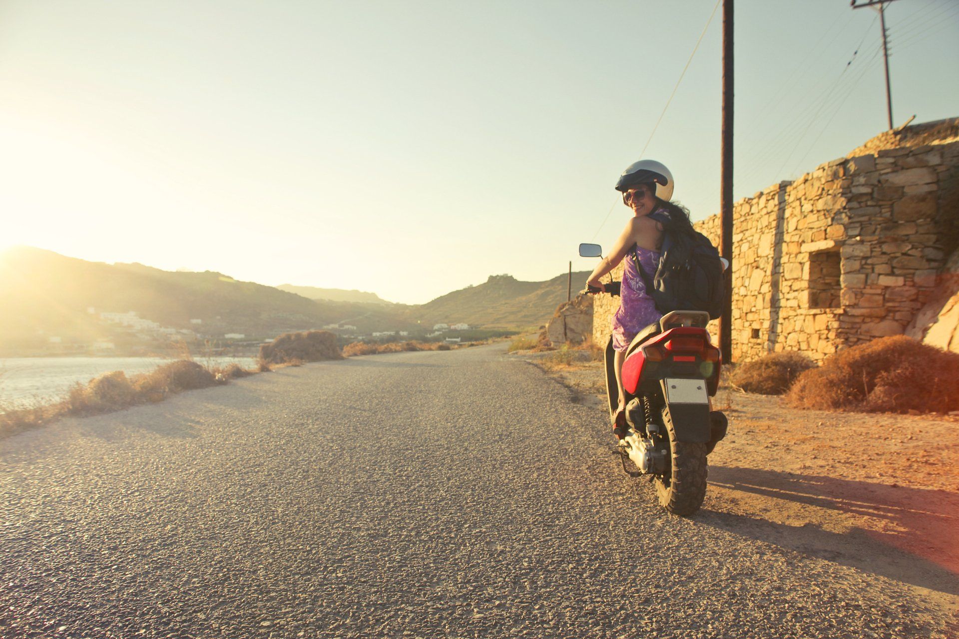 Eine Person mit Helm und Rucksack sitzt bei Sonnenuntergang auf einem Motorrad auf einer Küstenstraße auf der Insel Kreta.