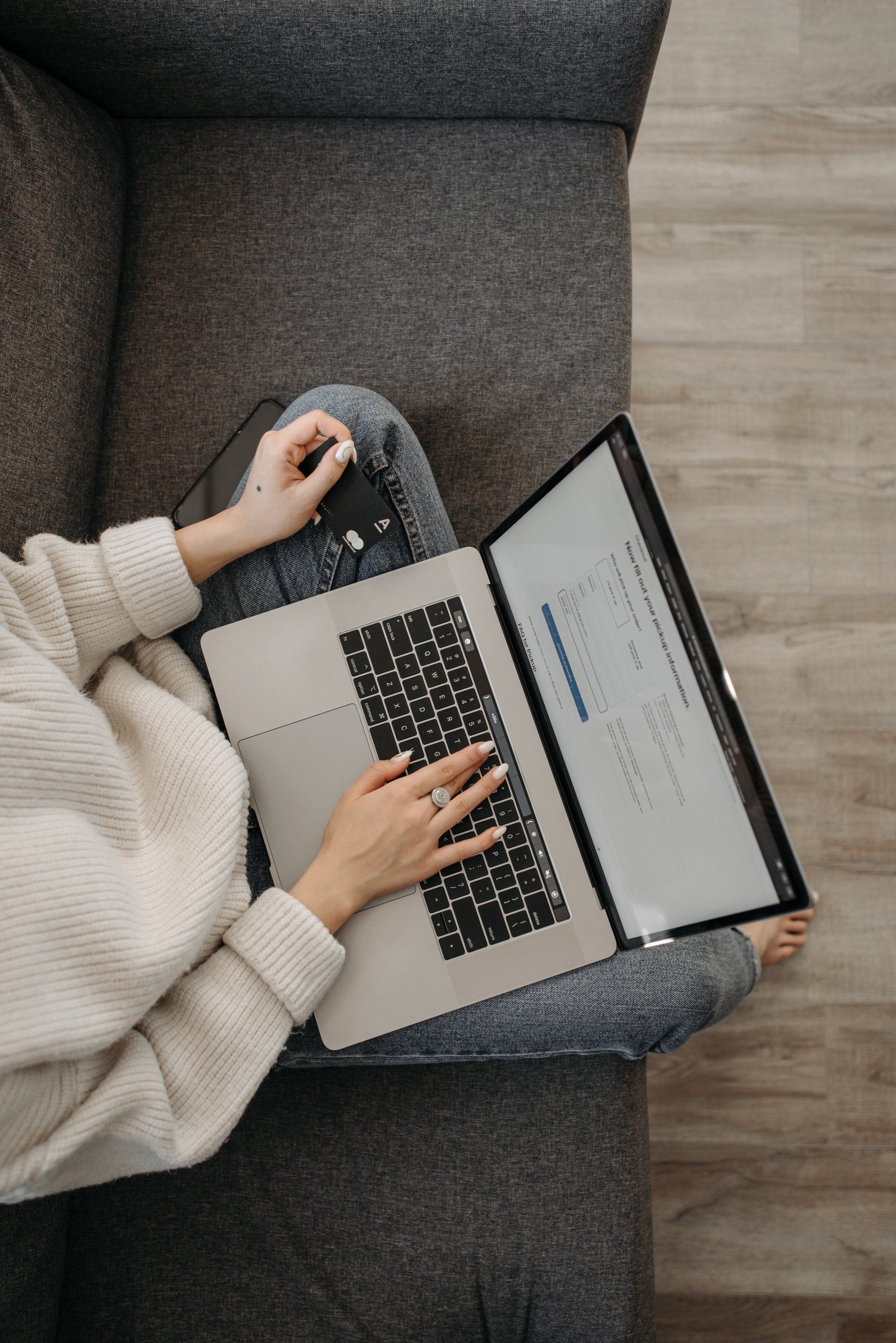 Person sitting on gray couch with laptop, typing. Wearing a white sweater and gray sweatpants.