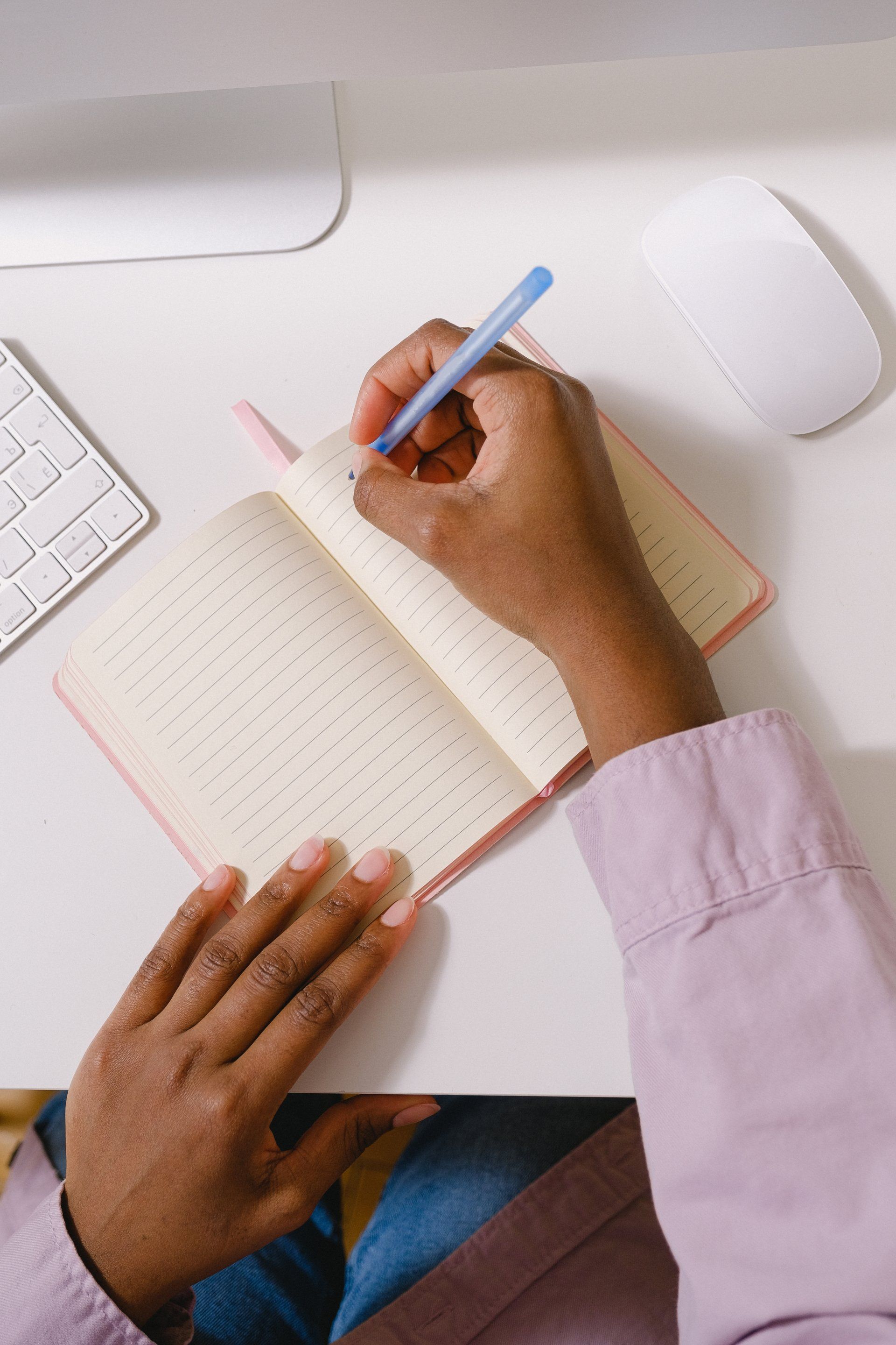 Person writing in a pink notebook with a blue pen, seated at a white desk with a keyboard and mouse.