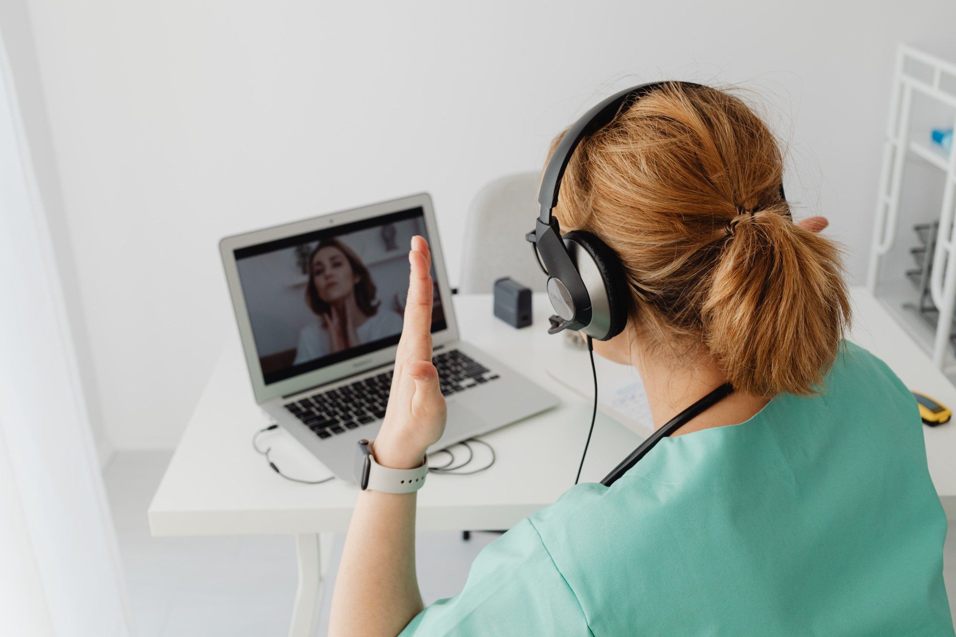 Person wearing headphones waves at a laptop during a video call in a bright white room