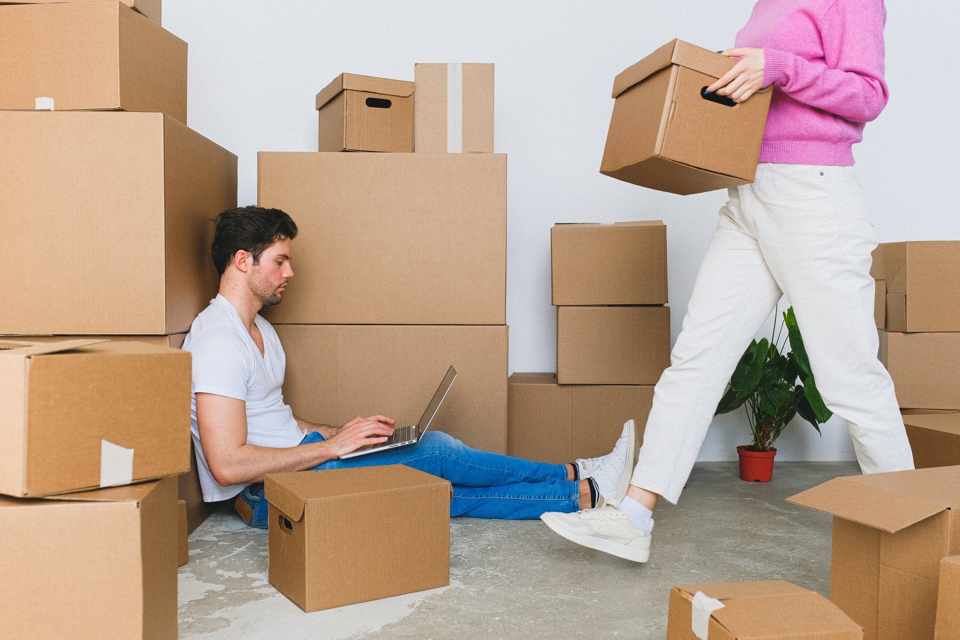 A man is sitting on the floor using a laptop while a woman is carrying boxes.