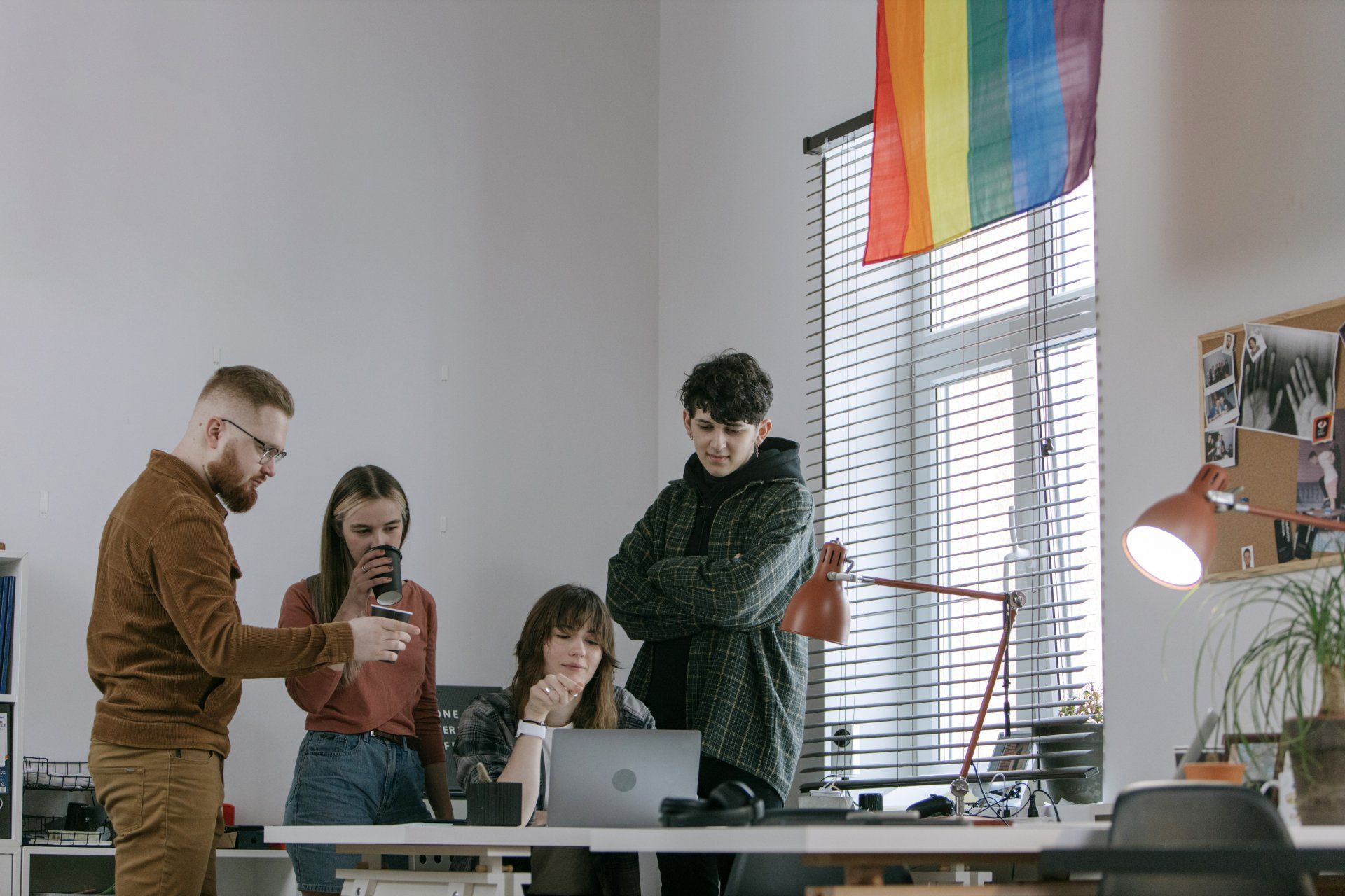 Four coworkers gather around a laptop in a bright office with a rainbow flag hanging by the window.