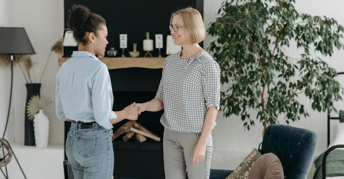 Two women shaking hands in a bright living room with a fireplace and plants.