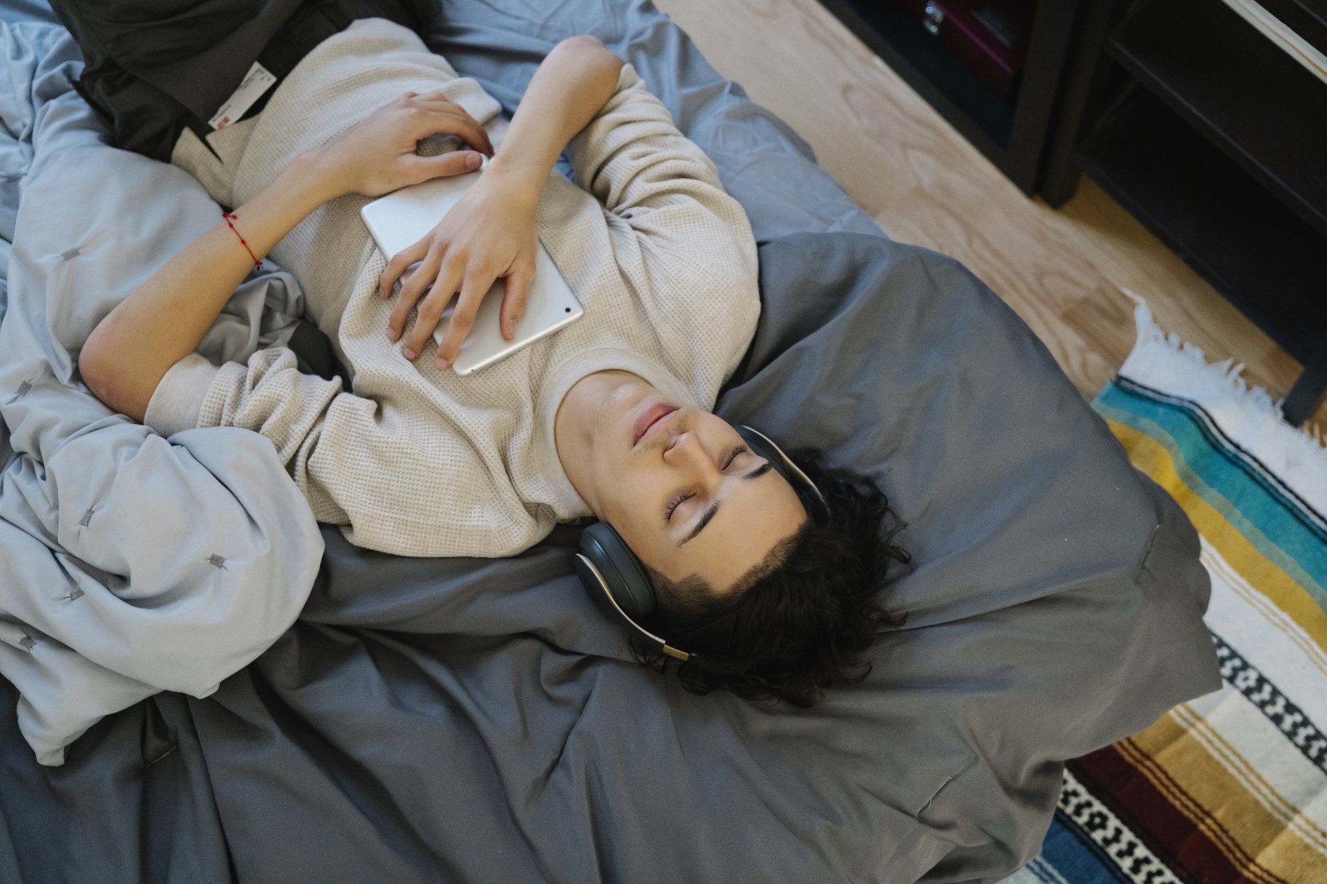 A man is laying on a bed wearing headphones and holding a tablet.
