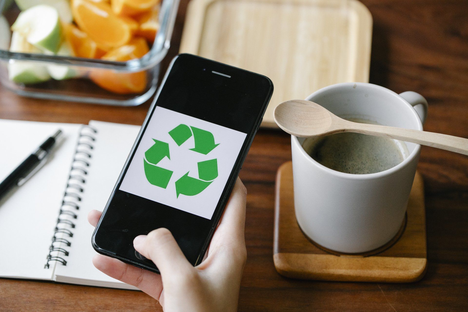 A person holds a smartphone displaying a green recycling symbol, with a notepad, fruit container, and coffee cup nearby.