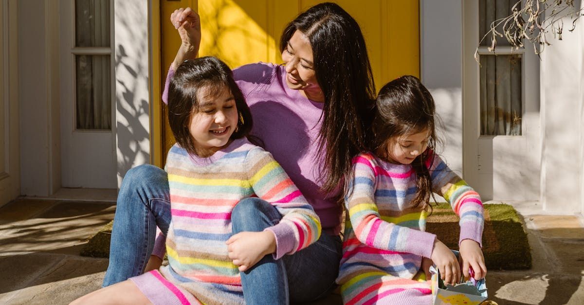 Two children in striped sweaters sitting on steps with an adult in purple behind them, in front of a house