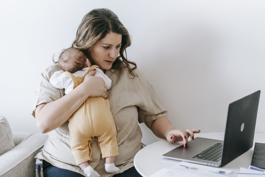 Woman holding a baby while using a laptop at a table in a bright room