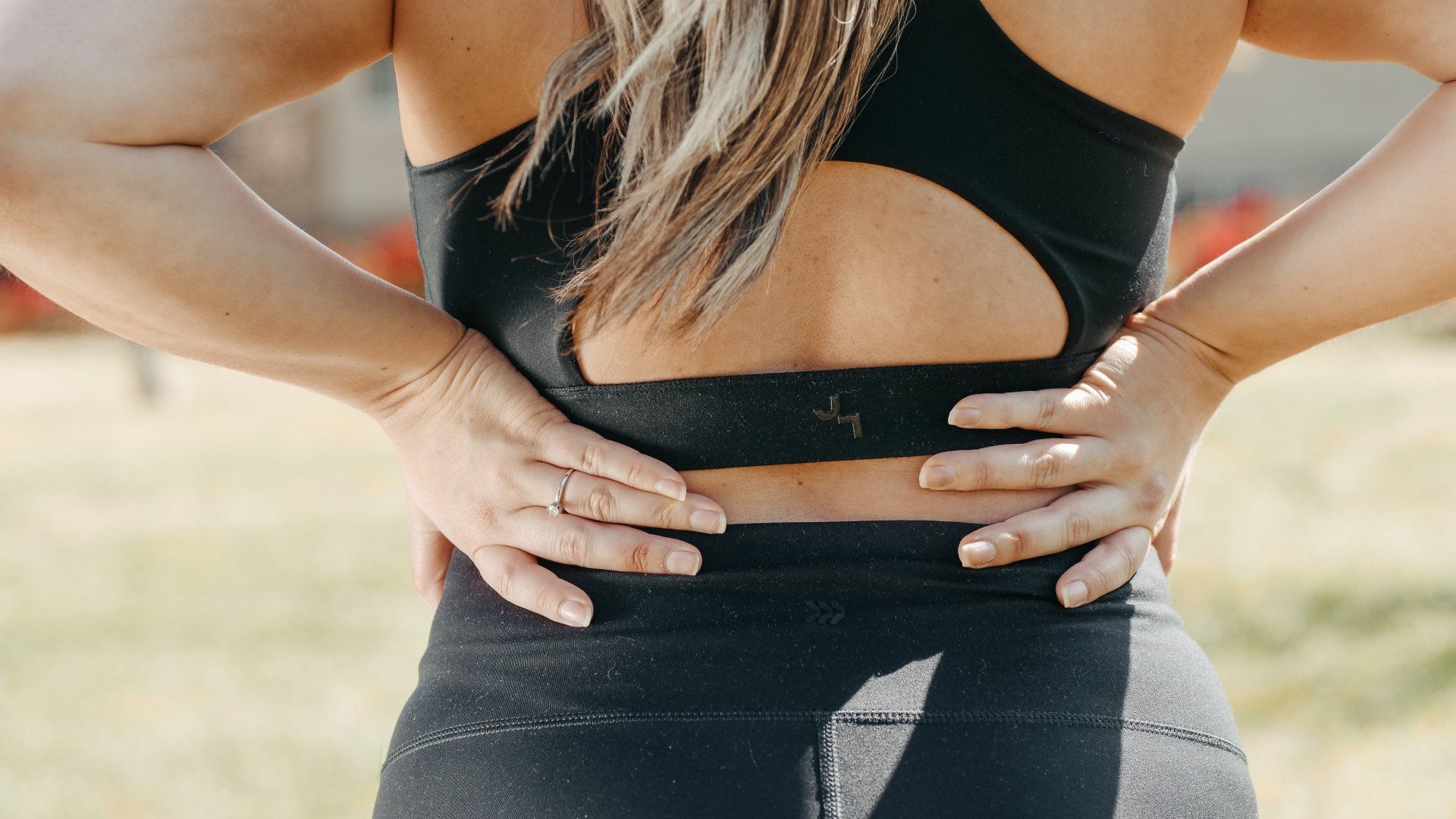 Woman in black athletic wear holding her lower back, outside in sunlight.
