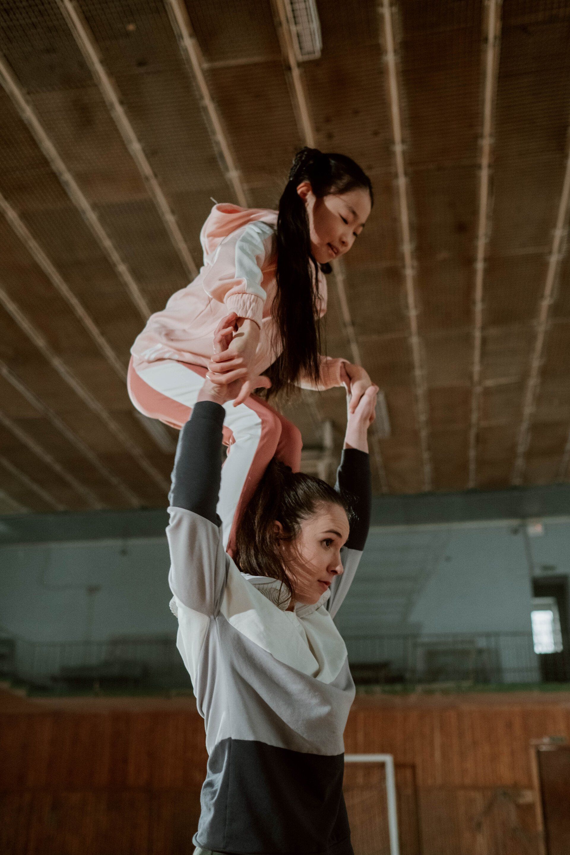 Twee mensen oefenen een gymnastiekact, waarbij de ene de andere boven zijn hoofd balanceert in een houten gymzaal.