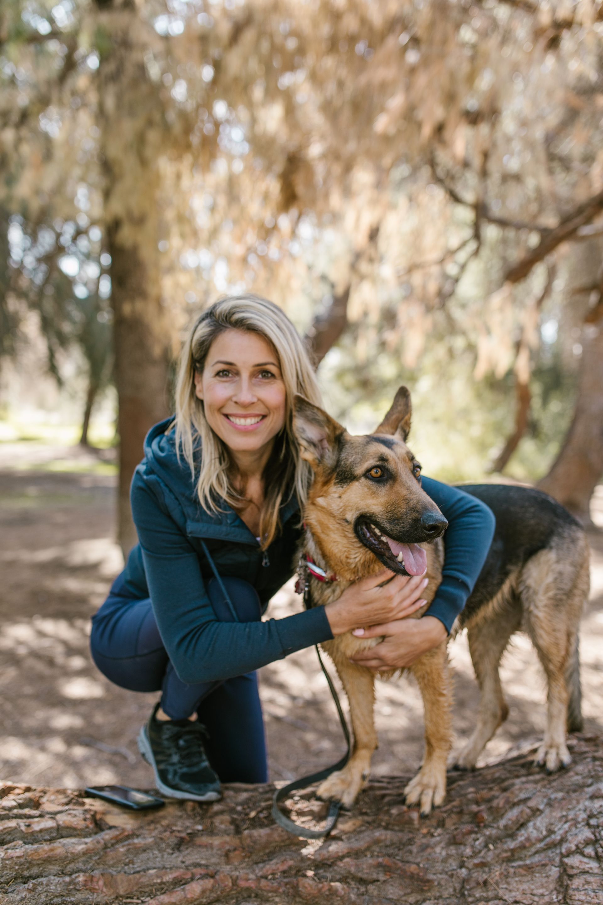 A woman is kneeling down next to a german shepherd dog in the woods.