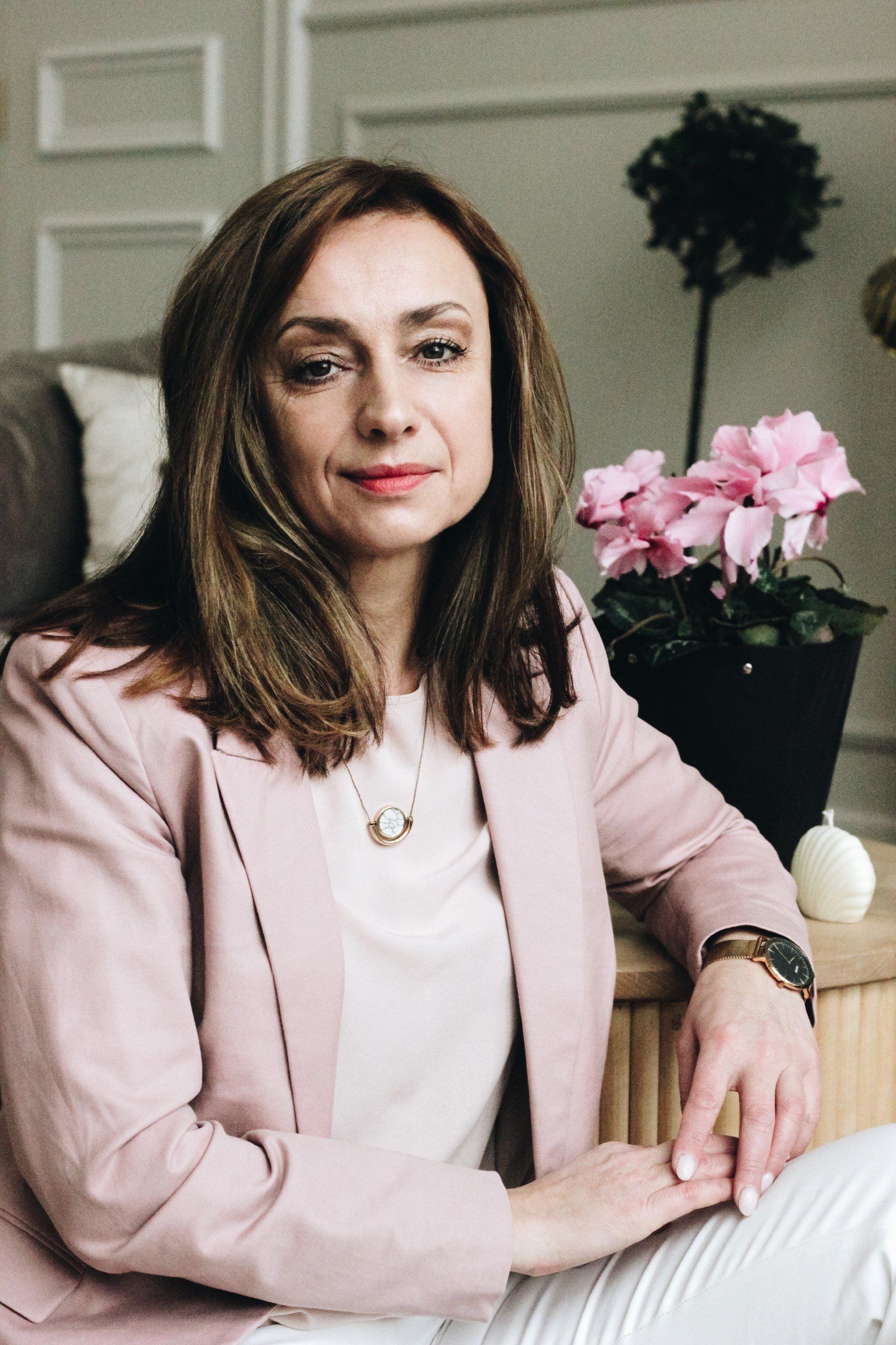 Woman in pink blazer smiles, seated indoors, near pink flowers, light background.