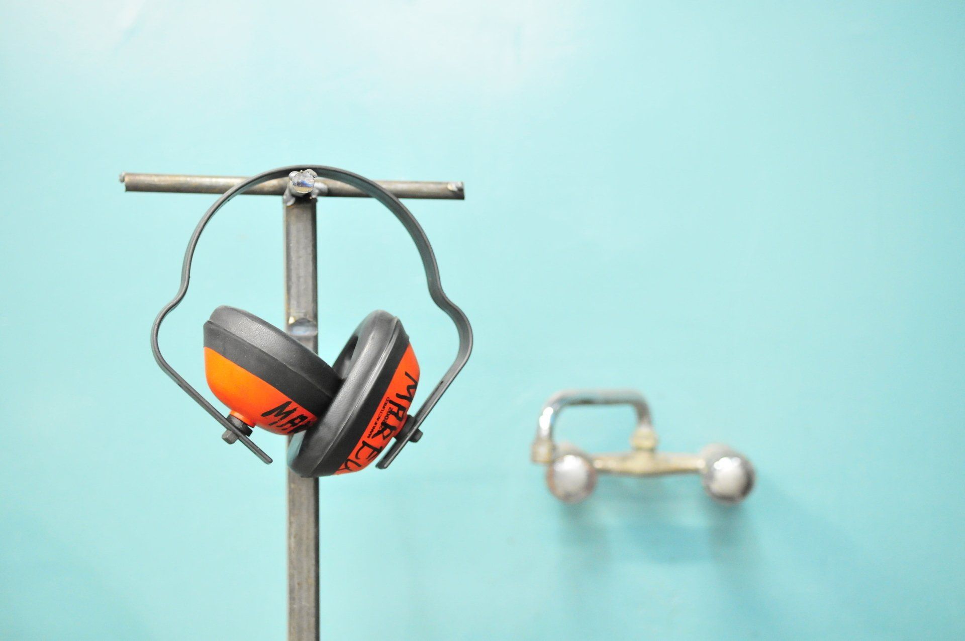 Orange and black earmuffs hanging on a metal stand against a turquoise wall; a blurred safety shower in the background.