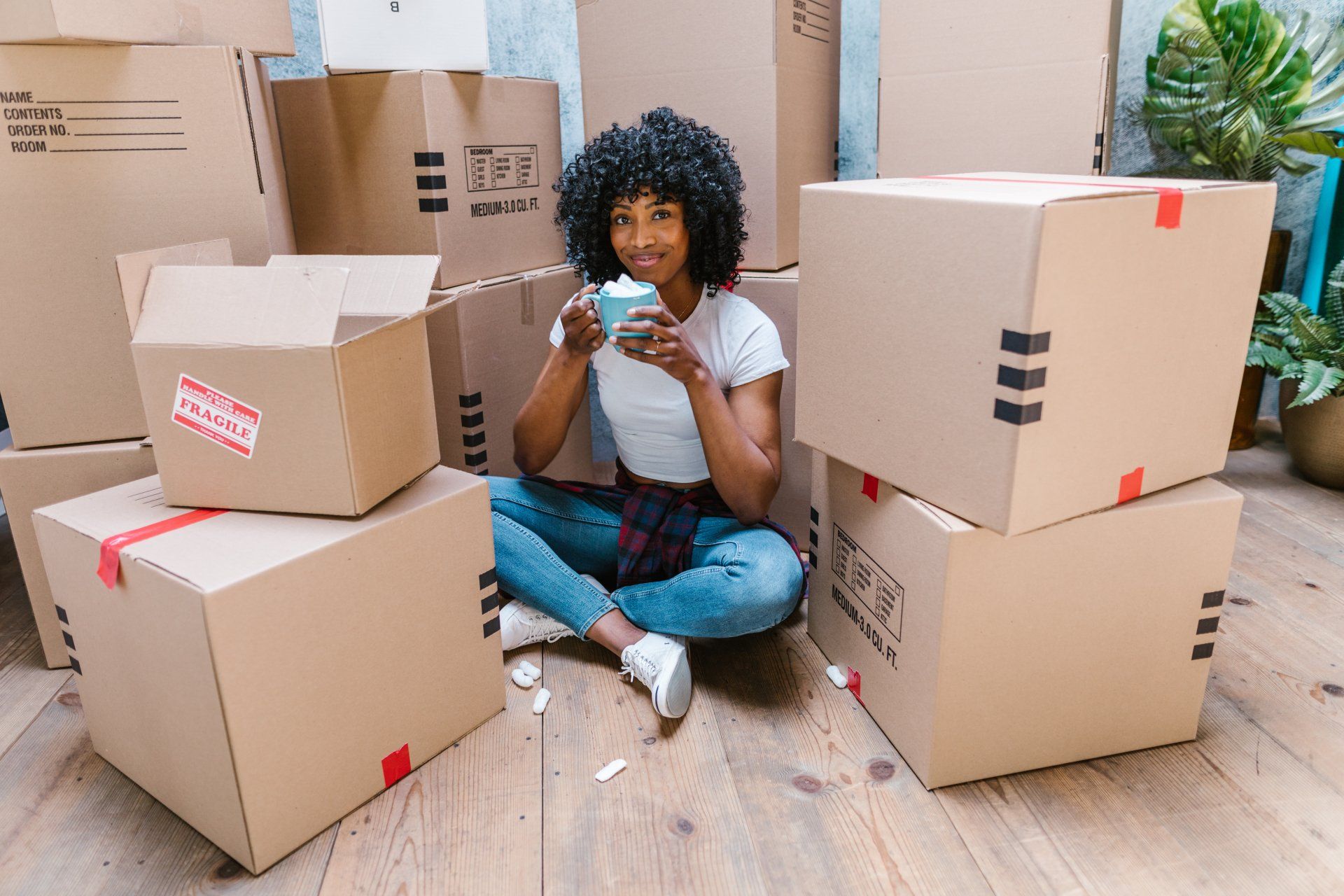 Woman sitting among moving boxes, smiling and holding a mug, indoors.