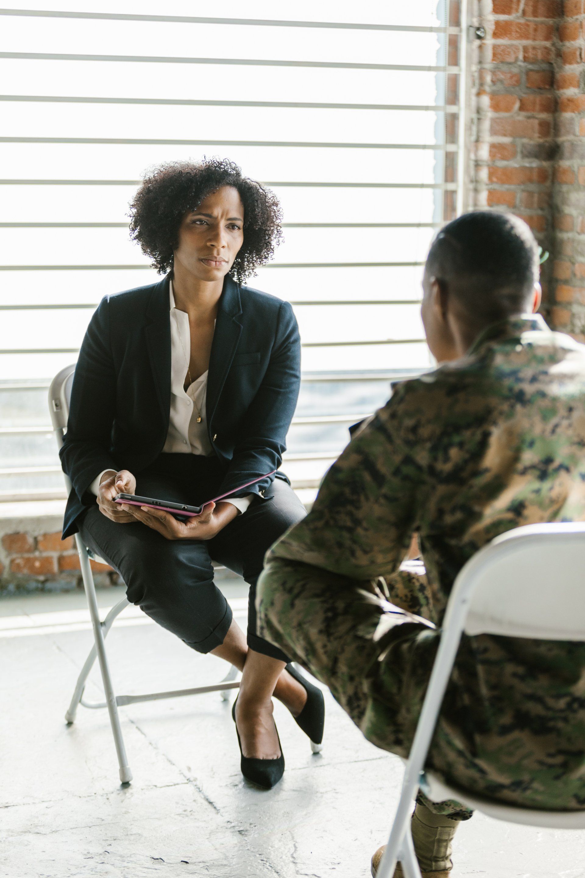 Woman in blazer and soldier in uniform sit facing each other in chairs, talking. Brick wall background.