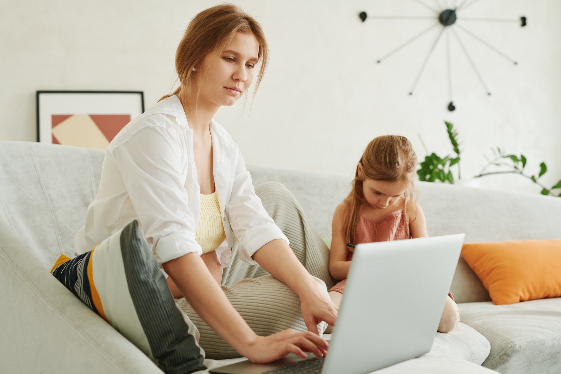 A woman is sitting on a couch using a laptop computer while a little girl looks on.