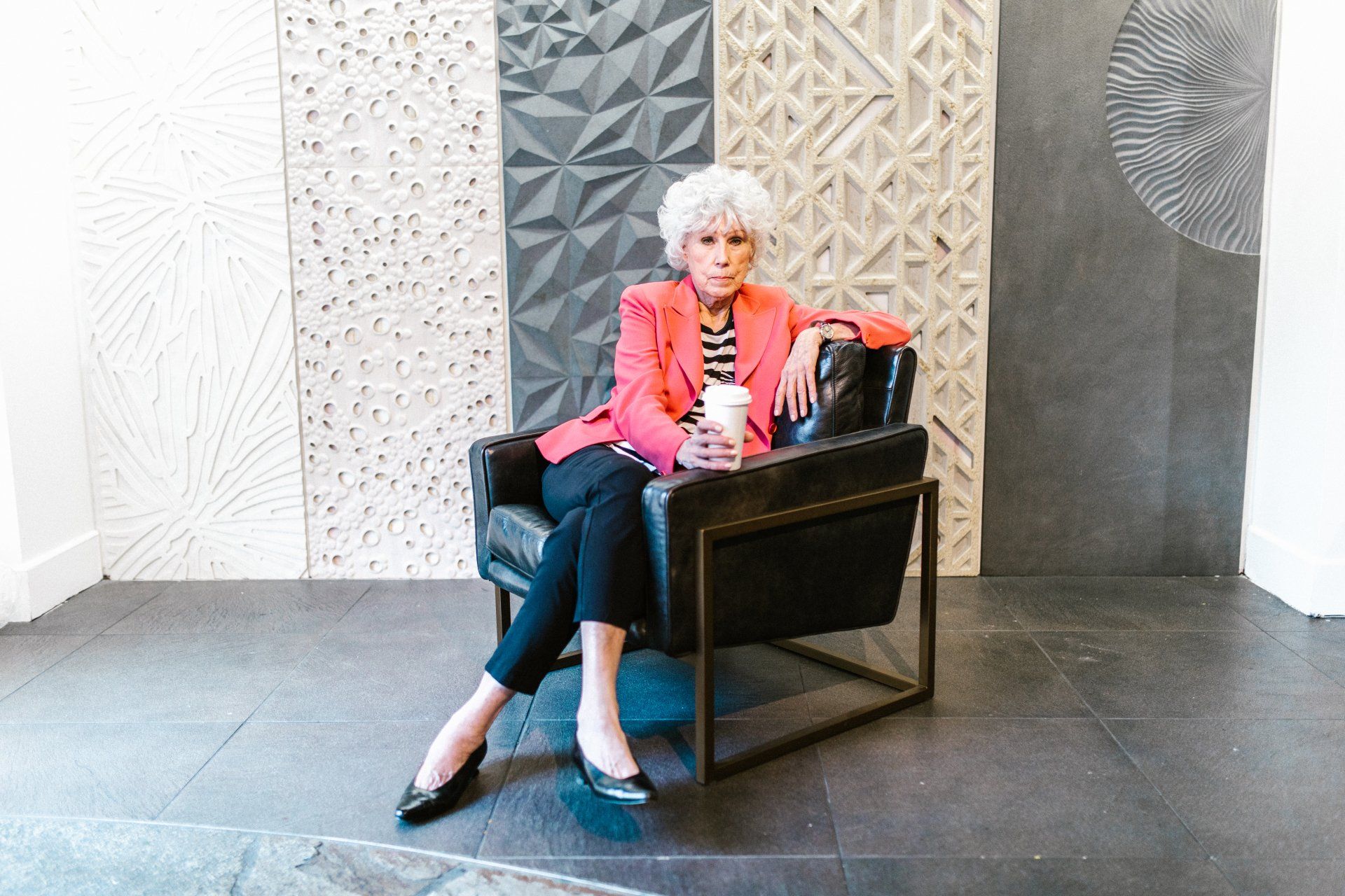 Woman in pink blazer and black pants sits in a chair, holding a coffee cup, with decorative wall panels behind her.