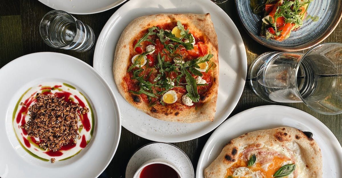 Overhead shot of a table with plates of food, including pizza, pasta, and appetizers. Glass of water visible.