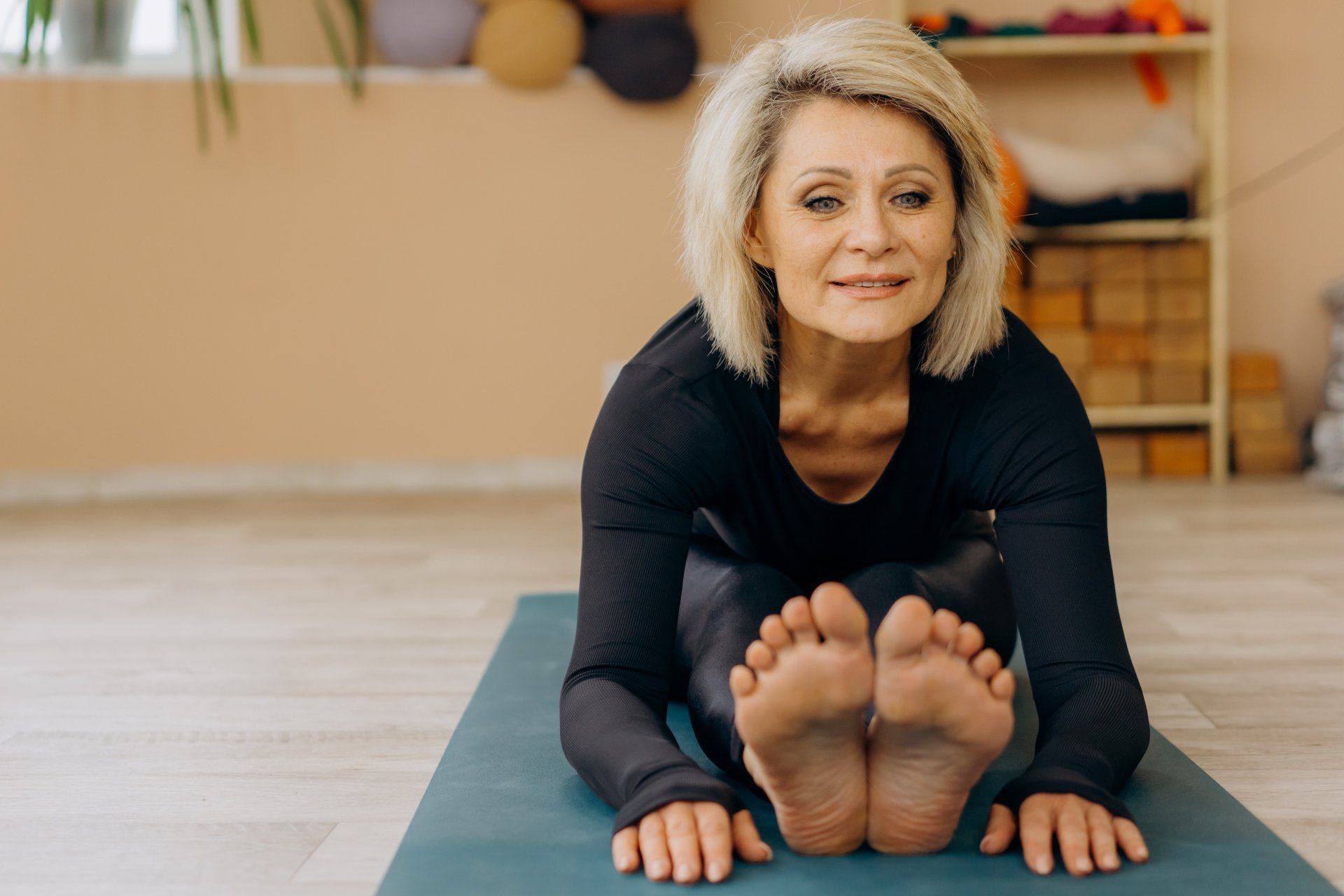 Mujer estirándose sobre una esterilla de yoga, con los pies extendidos. Interior con paredes beige.