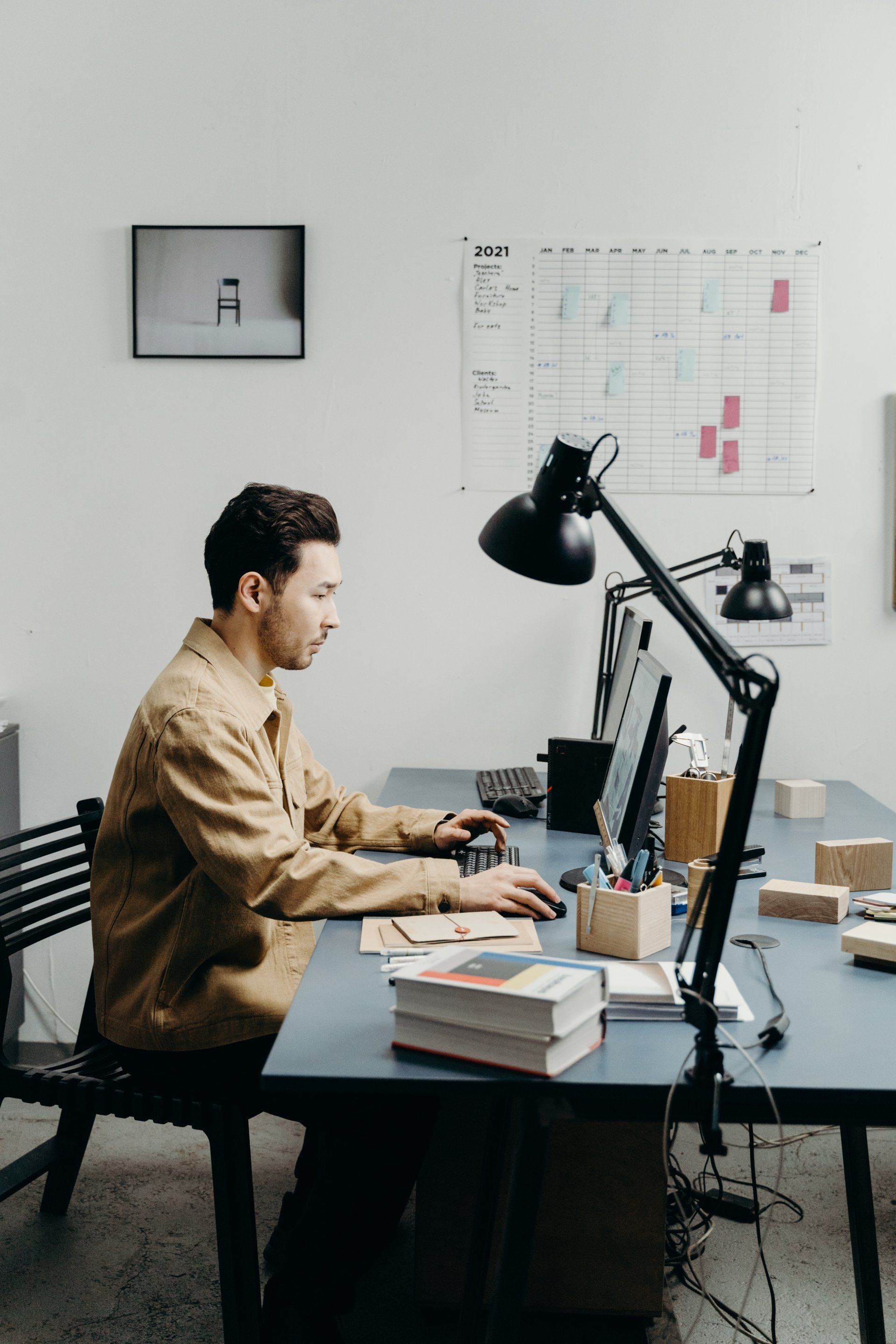 Un homme travaille à un bureau avec deux écrans et tape du texte. Bureau avec lampe, papiers et emploi du temps au mur.